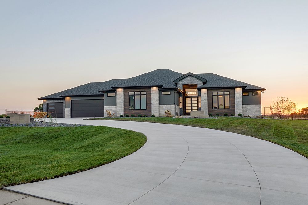 Modern gray house with dark roof, windows, and curved driveway on a grassy hill at sunset.