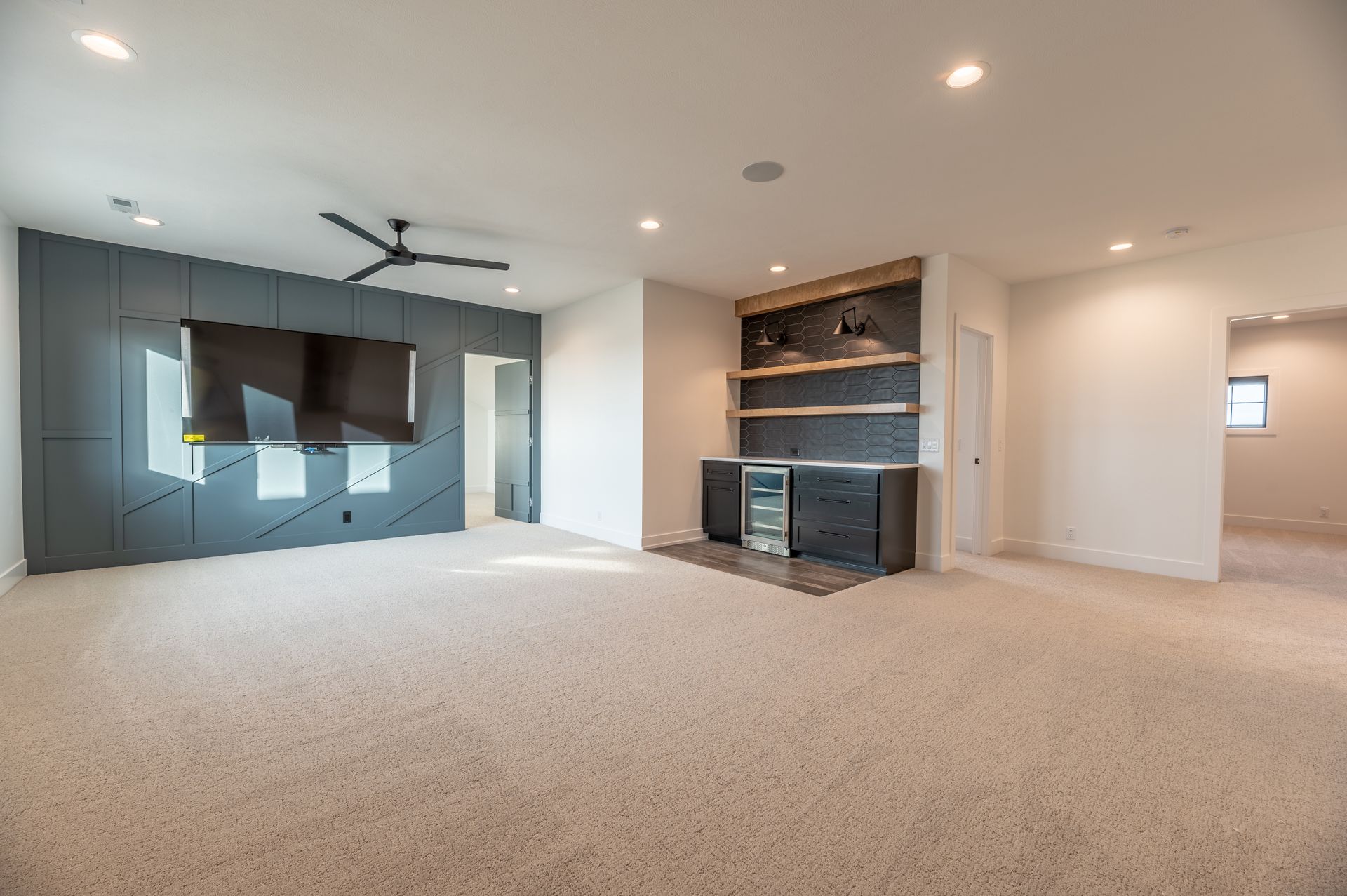 Empty, carpeted room with built-in bar and TV on accent wall. Recessed lighting and ceiling fan.