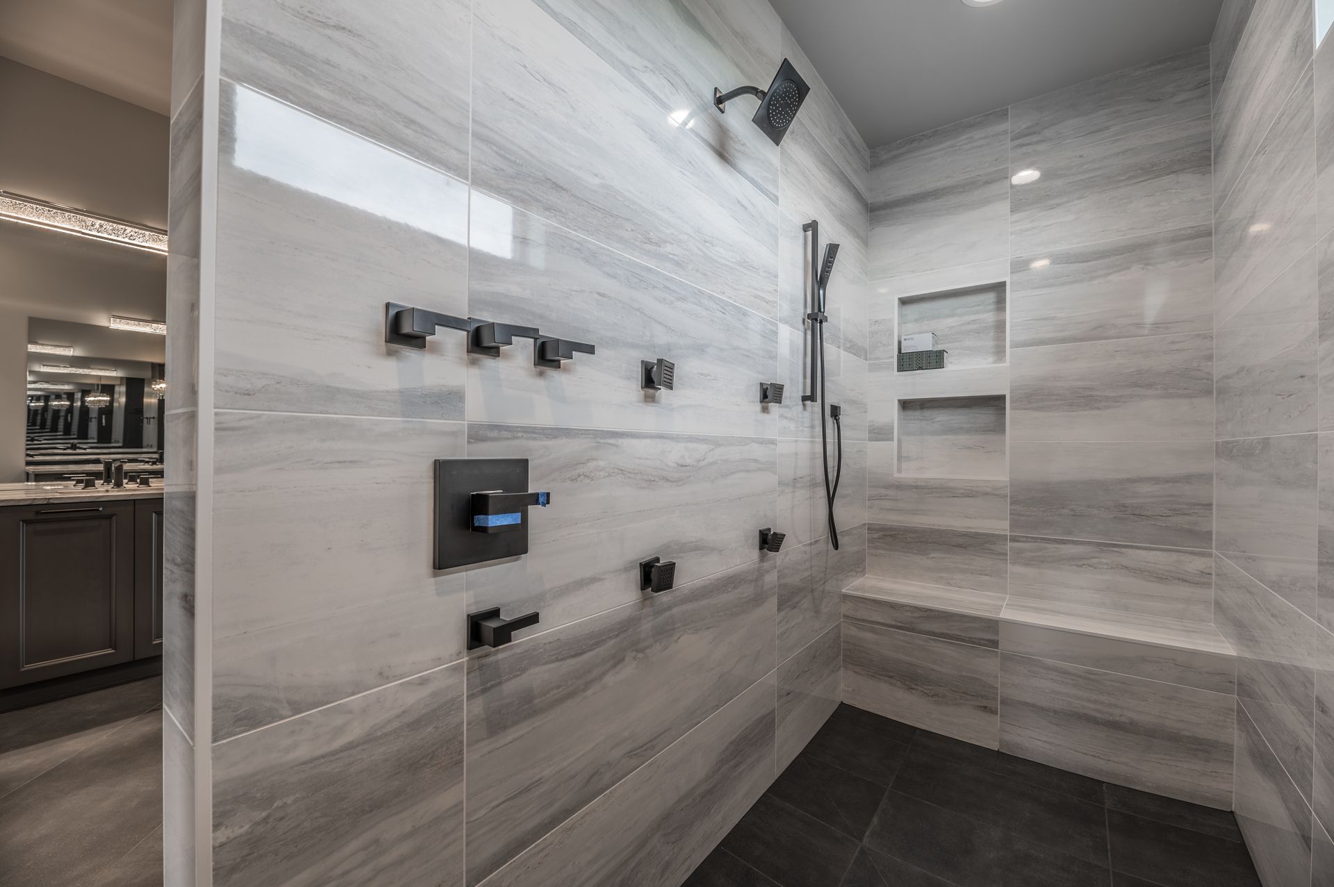 Modern bathroom with gray tiled walls, black fixtures, shower bench, and recessed storage.