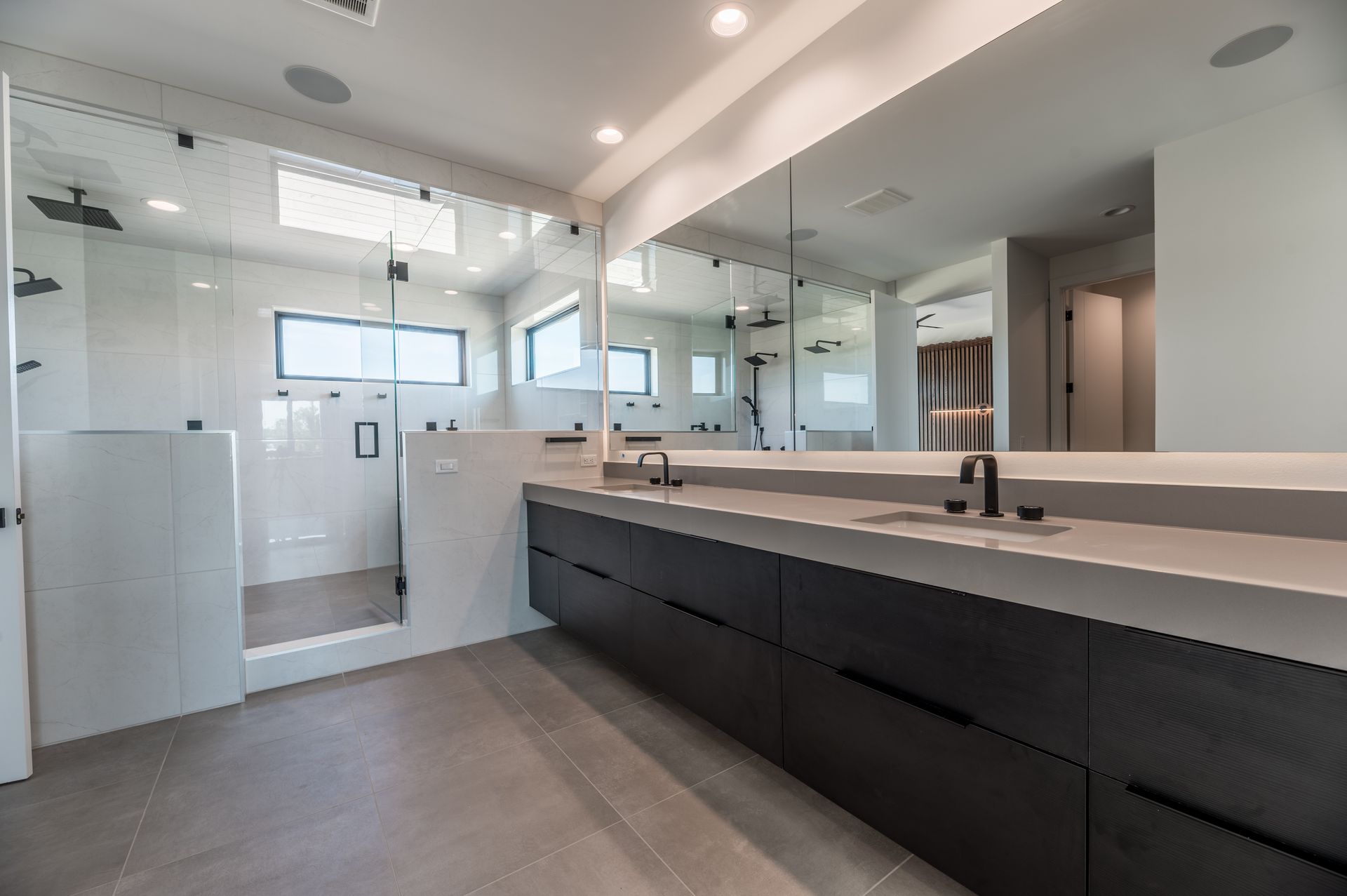 Modern bathroom with a large mirror, shower, and dark vanity. White walls and gray tile flooring.