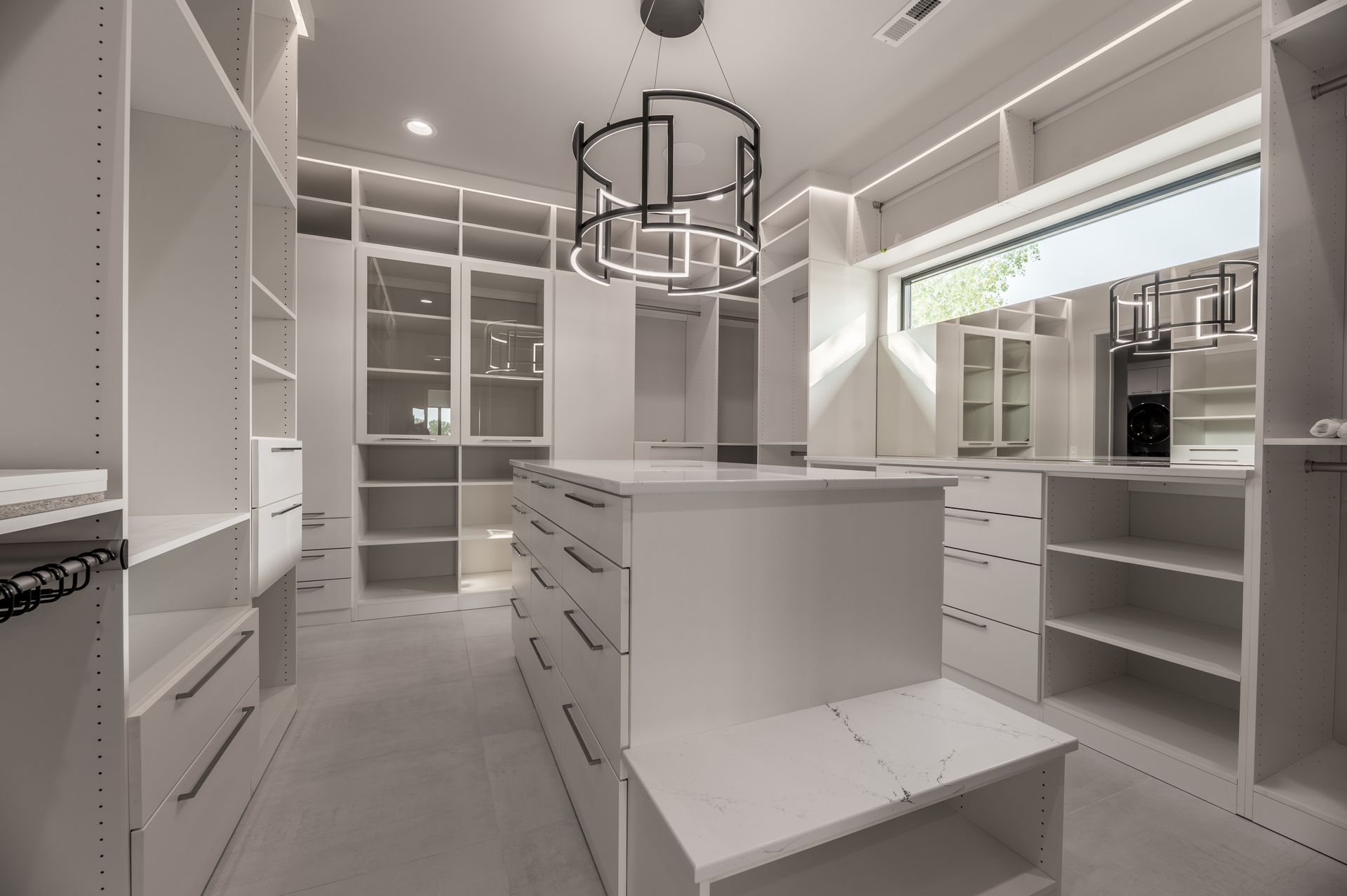 White walk-in closet with built-in shelving, drawers, and a central island. Modern chandelier hangs from ceiling.