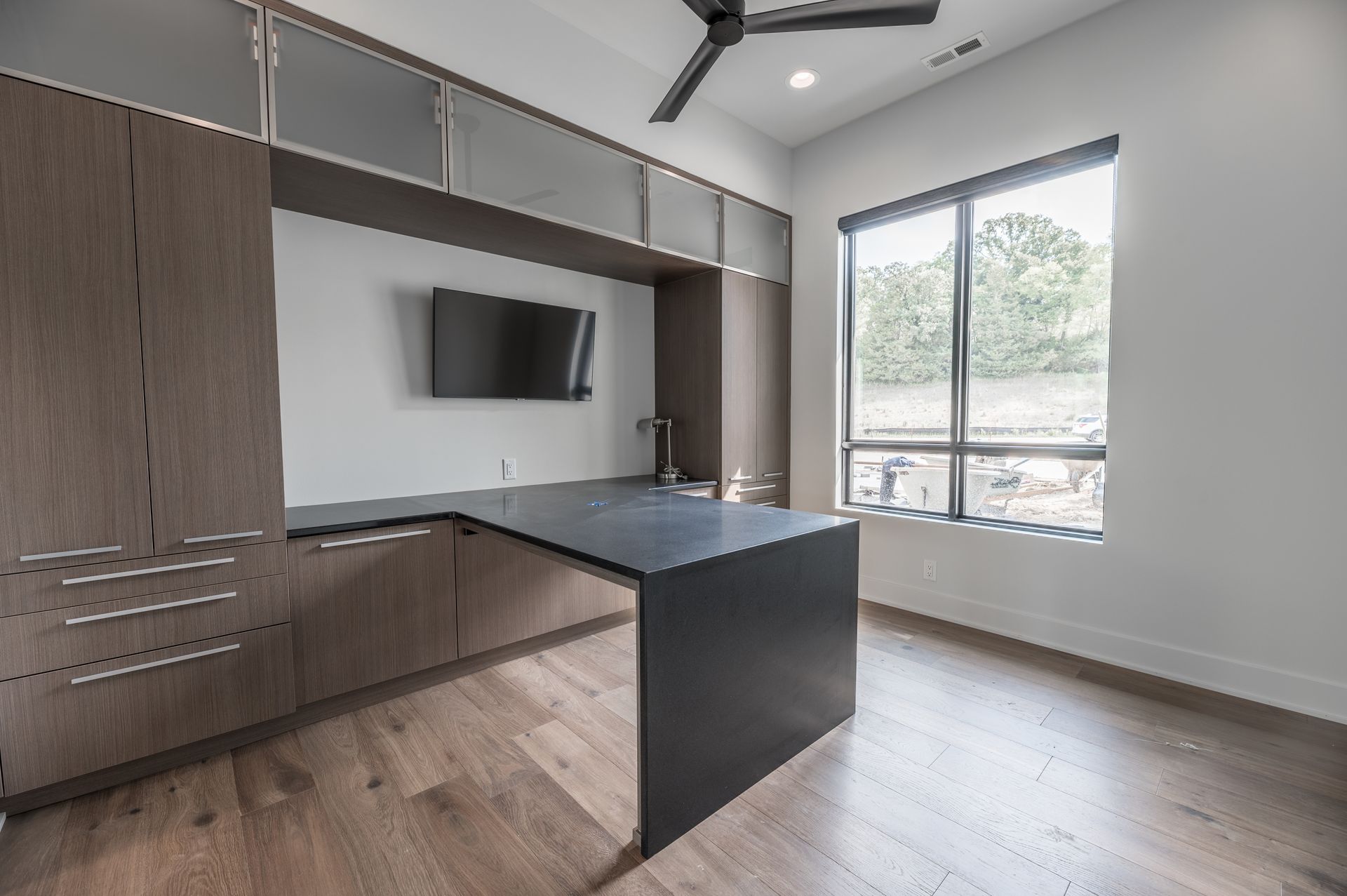Modern home office with a built-in dark wood desk and cabinetry, TV, and large window.