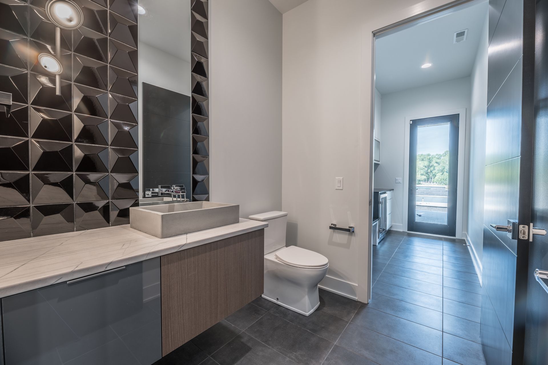 Modern bathroom with gray and wood vanity, black geometric accent wall, and open doorway.