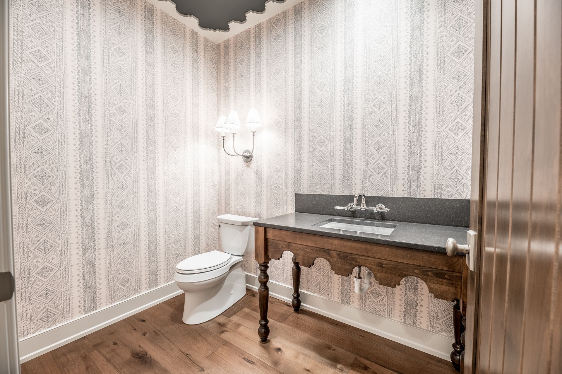 Small bathroom with patterned wallpaper, wooden floor, toilet, vanity with dark countertop, and decorative light fixture.
