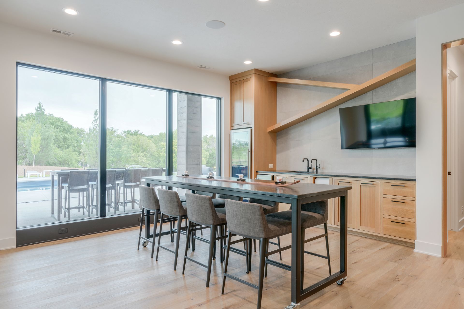 Modern bar area with large windows overlooking a pool, bar stools, counter with sink, and a mounted TV.