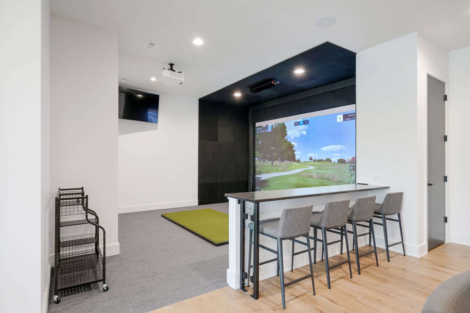 Golf simulator room with screen, bar seating, projector, and golf mat.