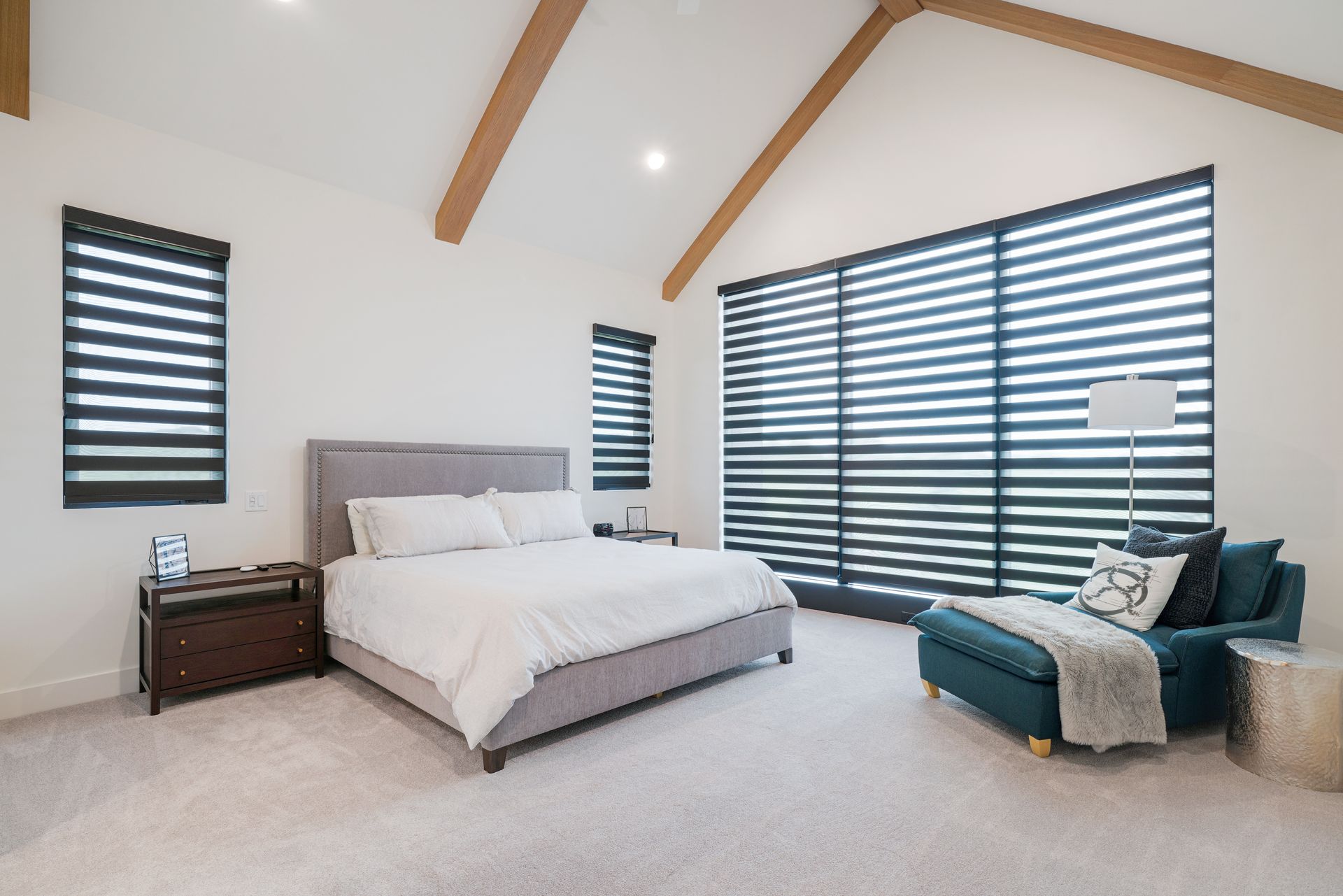 Bedroom with a bed, chaise lounge, and blinds. Beige carpet, white walls, and wood beams.