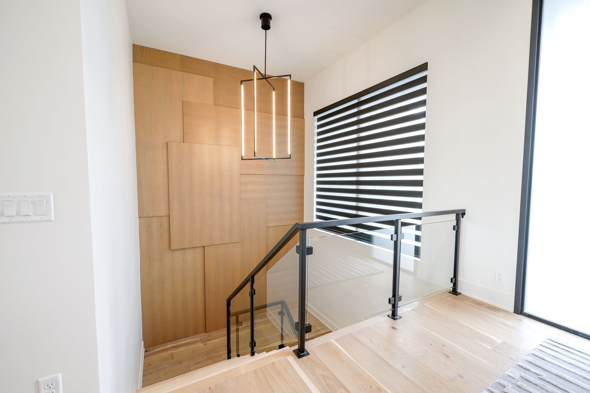 Stairwell with light wood paneling, black railing, and modern light fixture. Window with striped blinds.