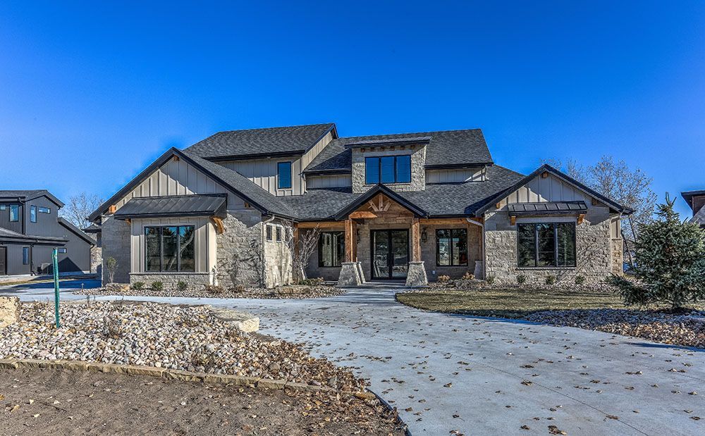 Stone-clad, two-story house with a driveway. Wooden accents around windows and entrance against a clear blue sky.