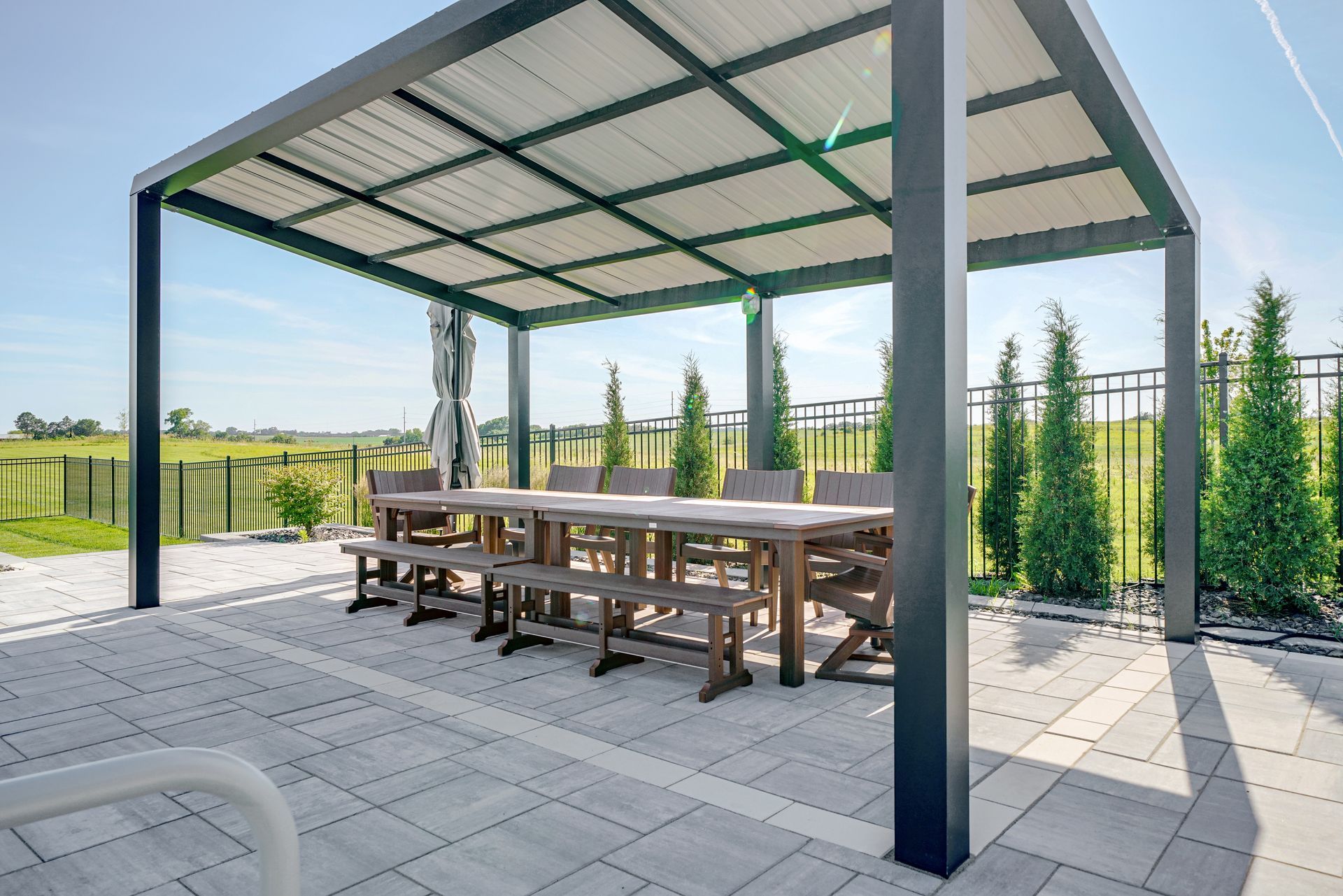 Pergola with long wooden table and benches on a patio. Green field and trees in the background.