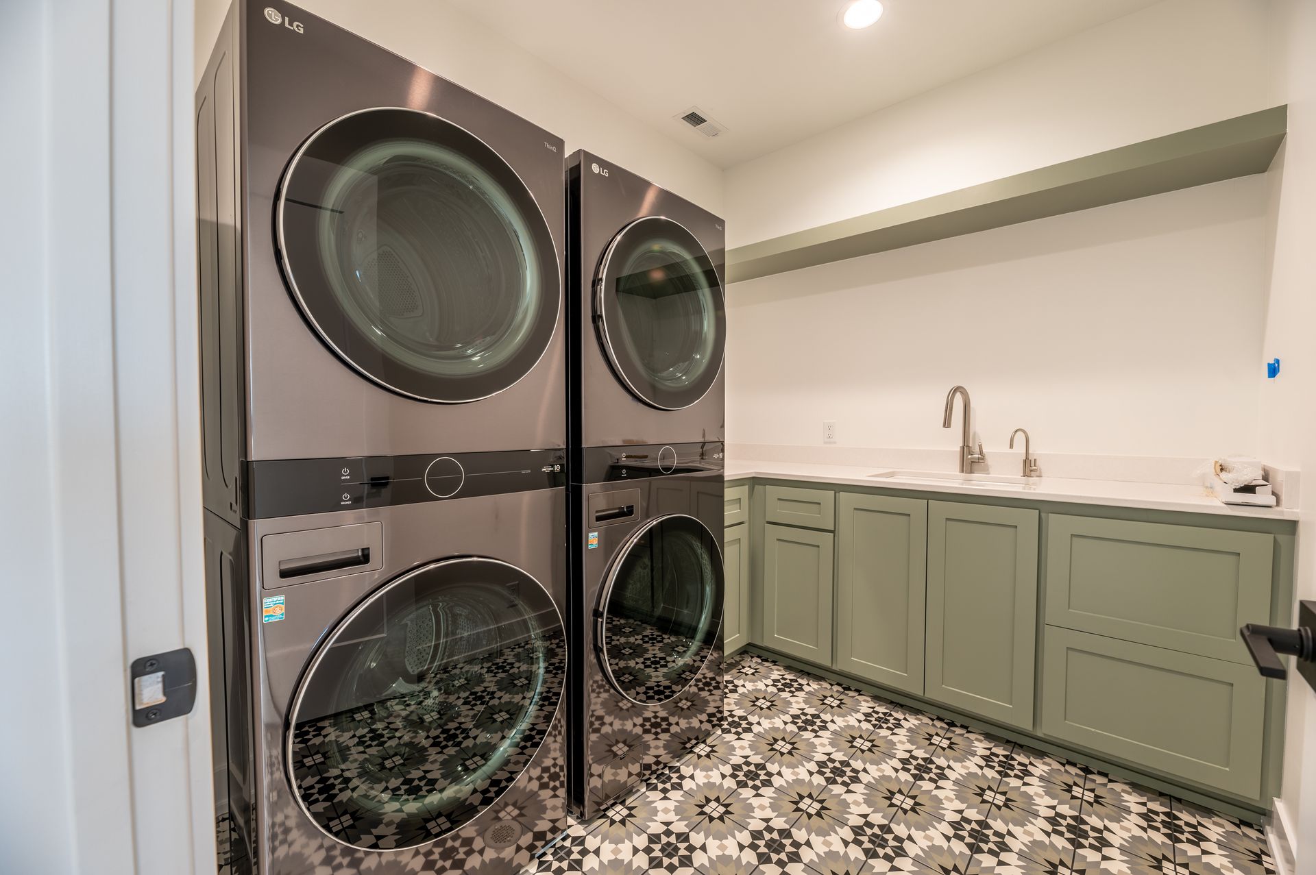 Laundry room with stacked washer/dryer, sink, green cabinets, and patterned tile floor.