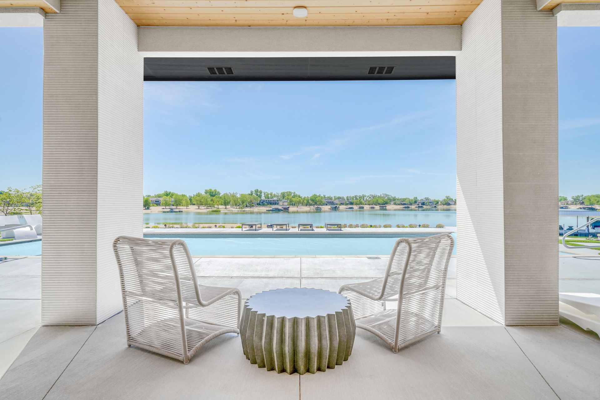 Two white woven chairs and a table on a patio, overlooking a pool and waterfront.