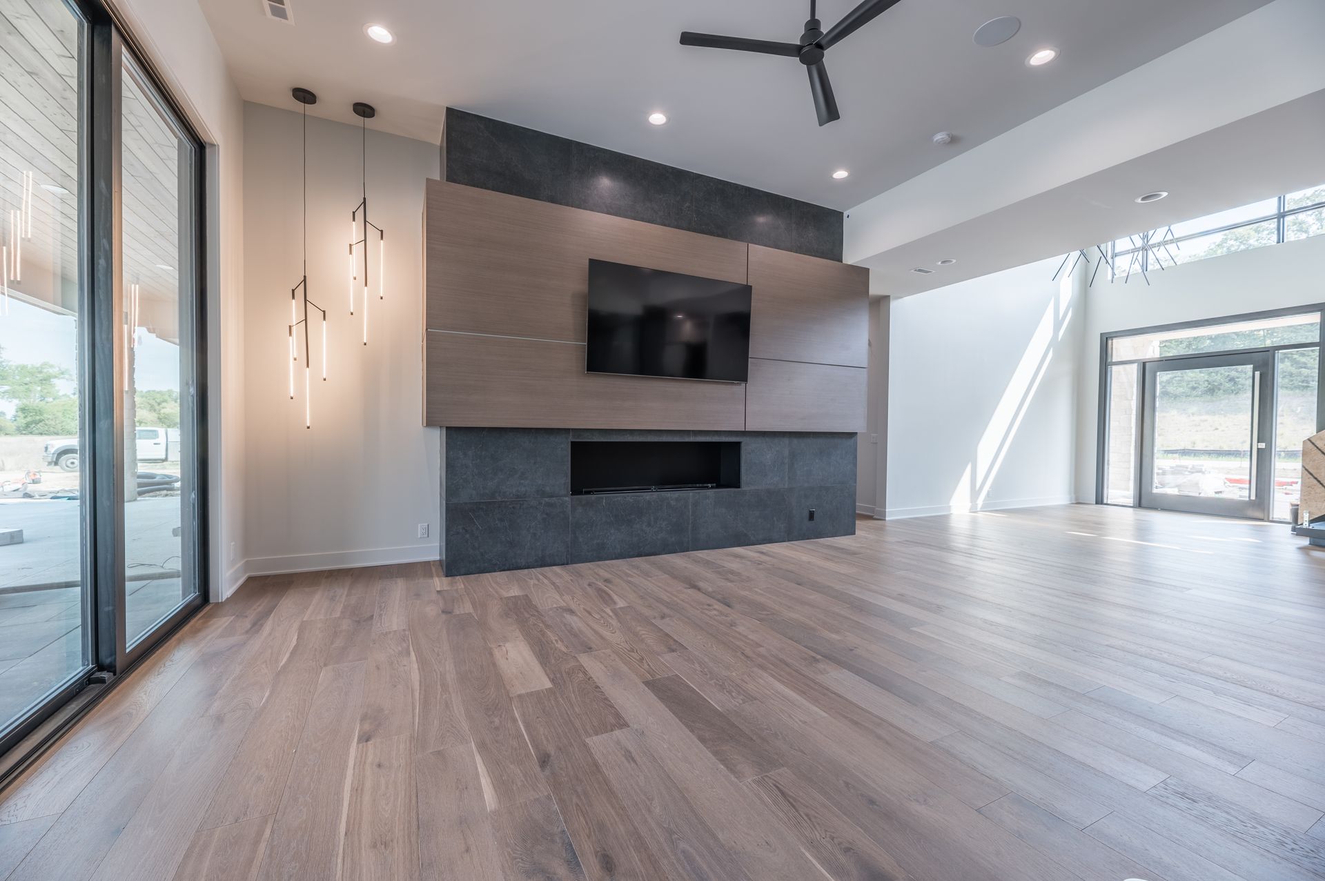 Modern living room with a fireplace, TV, and large windows. Wooden floors, gray and tan accents, and minimalist lighting.