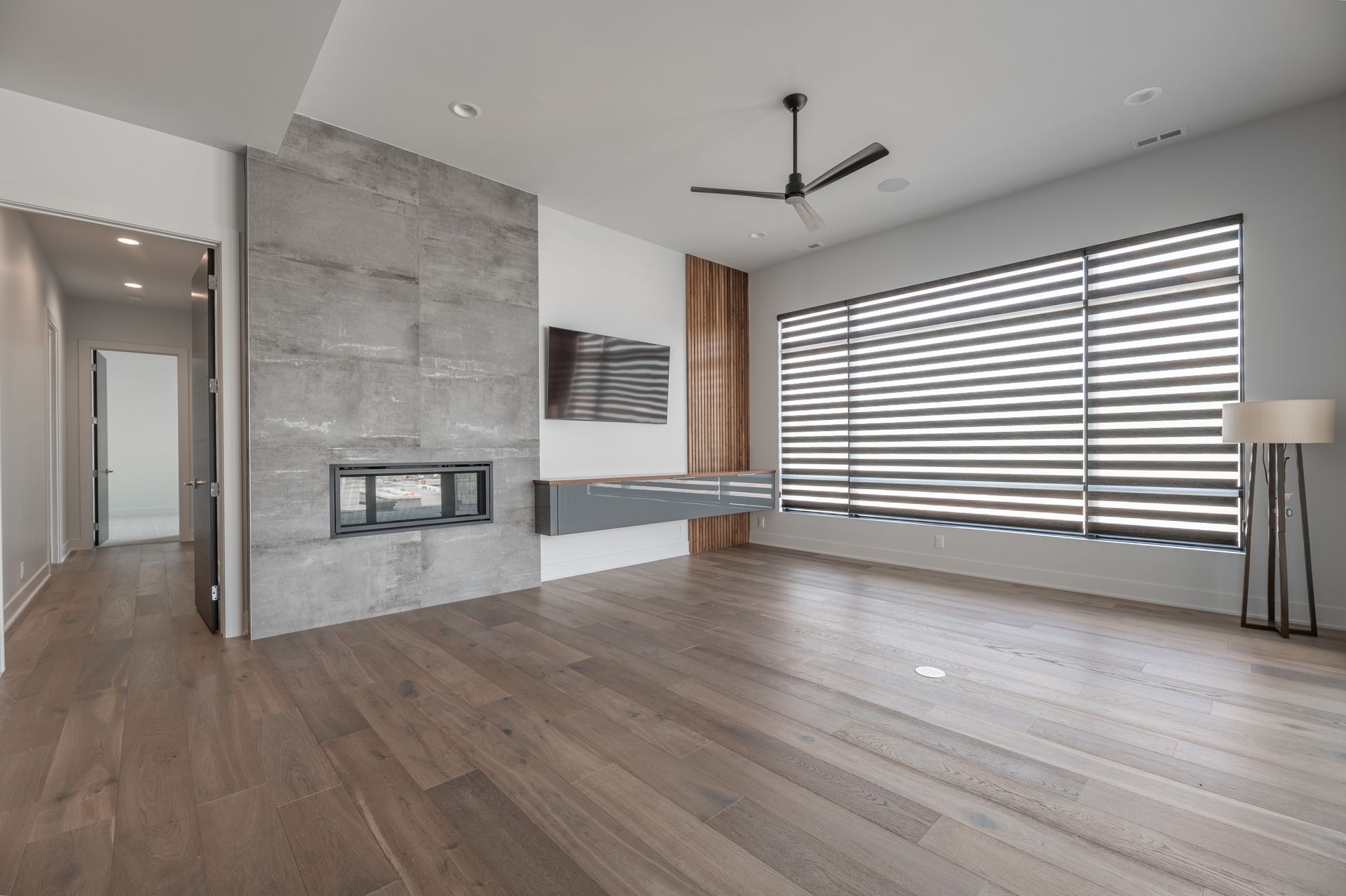 Modern living room with fireplace, hardwood floors, large window with blinds, and ceiling fan.