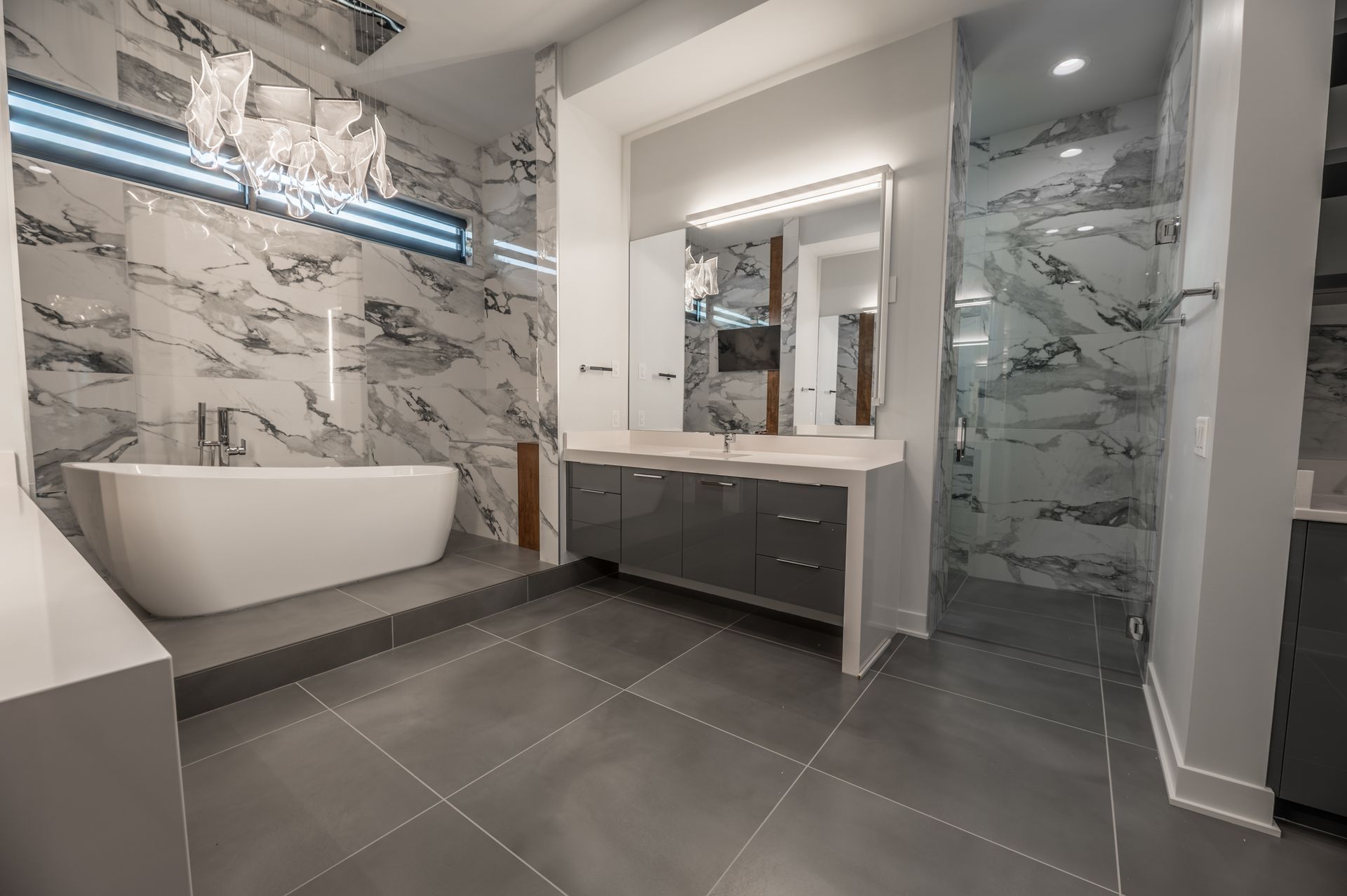 Modern bathroom with marble walls, freestanding tub, glass shower, gray cabinetry, and floor.