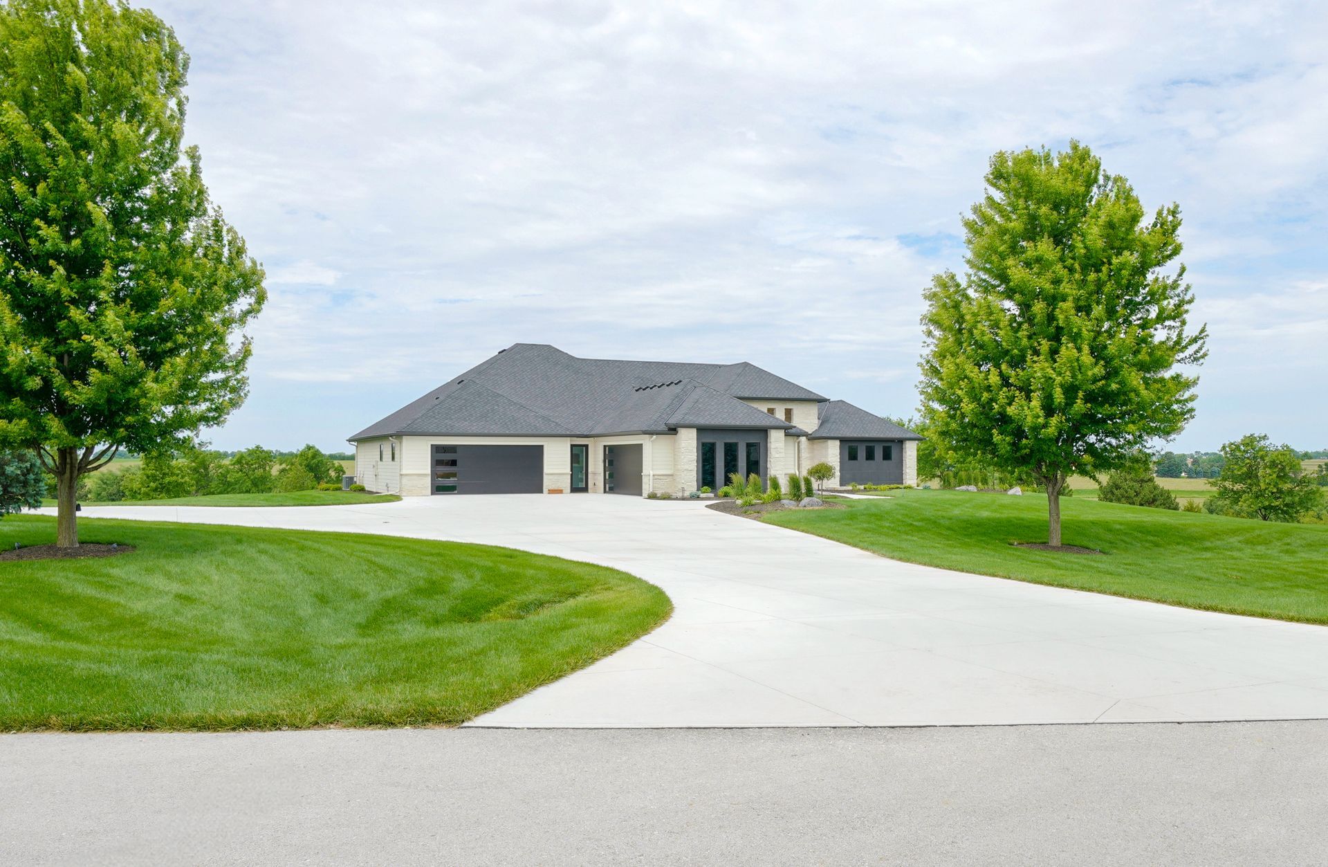 Modern house with a gray roof, light exterior, and long concrete driveway, flanked by green lawns and trees.