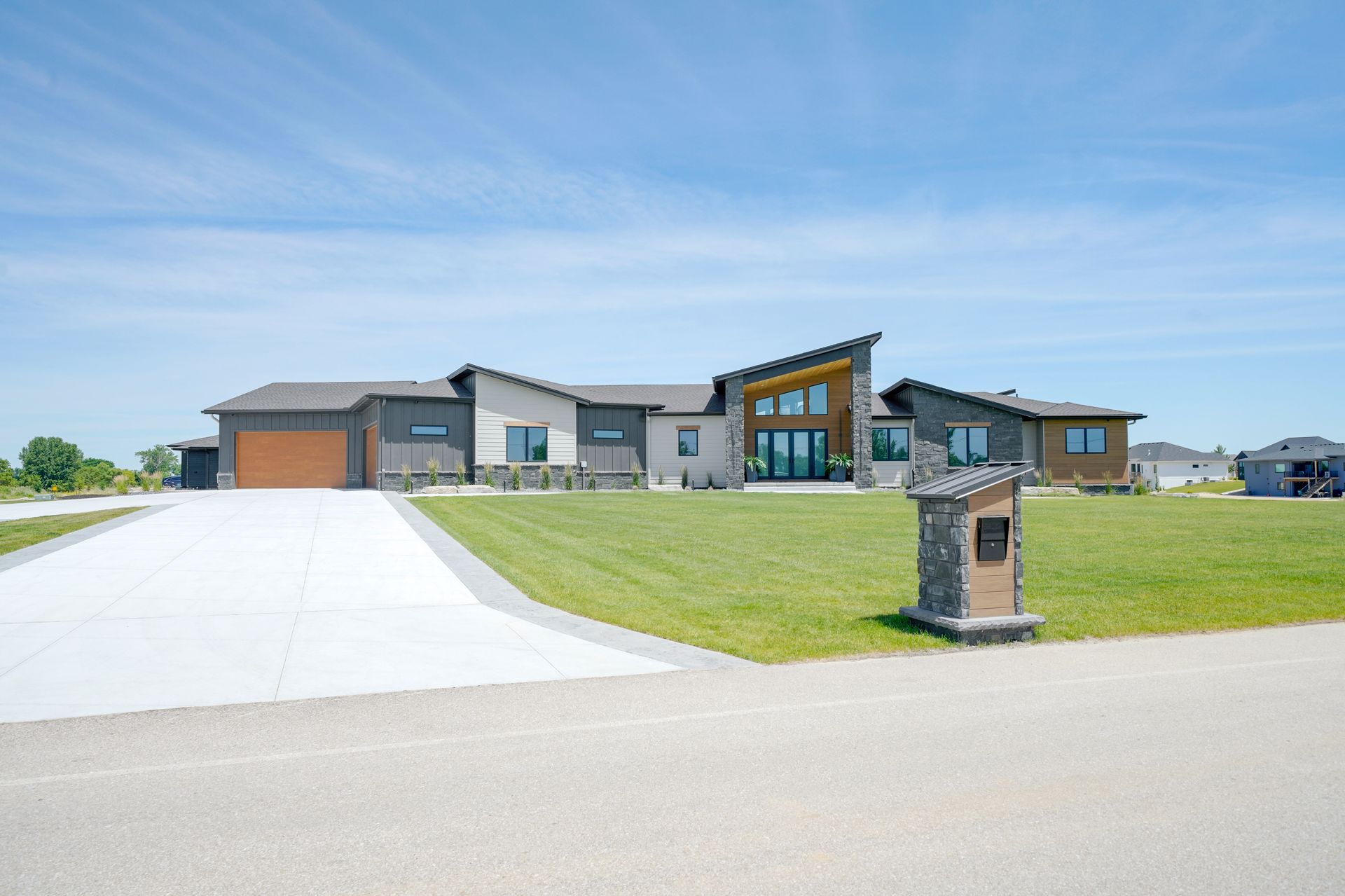 Modern house with gray and brown exterior, large windows, and a wide driveway on a sunny day.