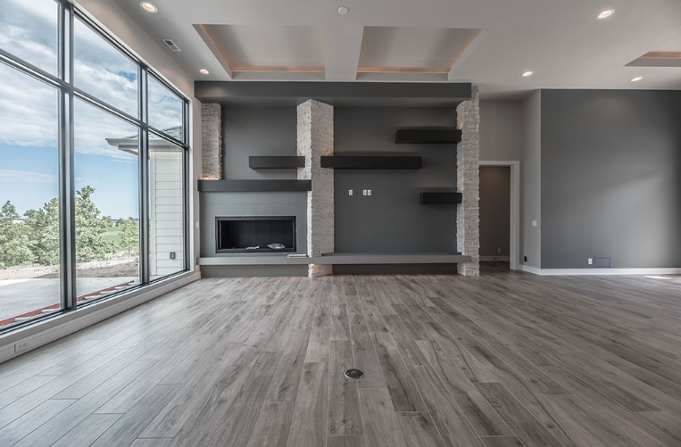 Empty modern living room with gray walls, fireplace, large windows, and wood flooring.