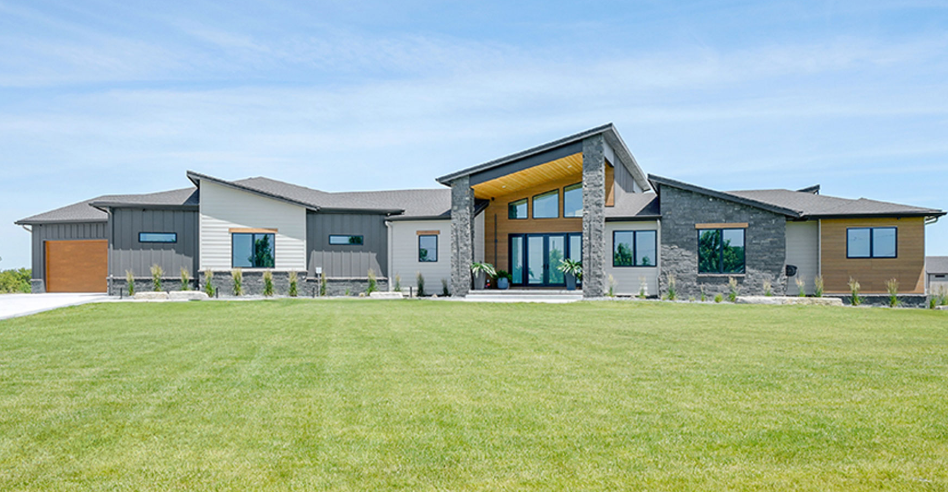 Modern one-story house with a long, grassy yard under a blue sky. Exterior features stone, wood, and glass elements.