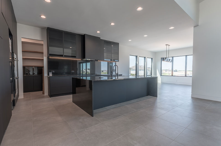 Modern kitchen with dark gray cabinets, island, and tile floor; white walls and large windows.