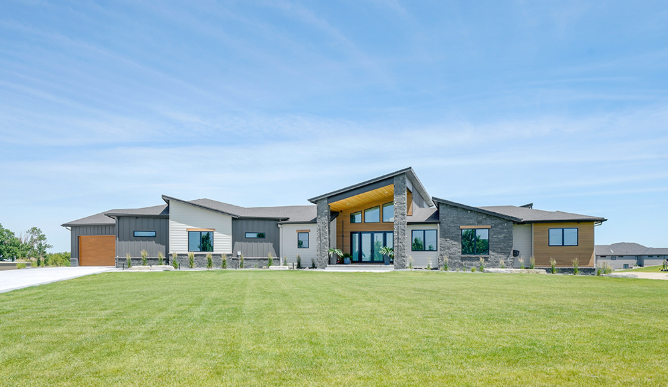 Modern one-story house with varied siding colors under a blue sky, fronted by a large lawn.