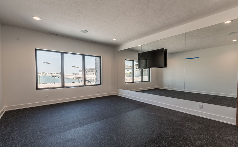 Empty exercise room with black flooring, a large mirror, windows, and a mounted TV.