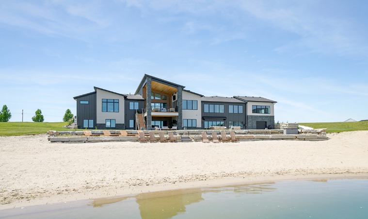 Modern house with gray and black exterior on a sandy beach by water under a blue sky.