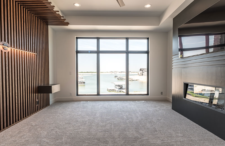 Empty room with large window overlooking water. Wooden accent wall and gray carpet.