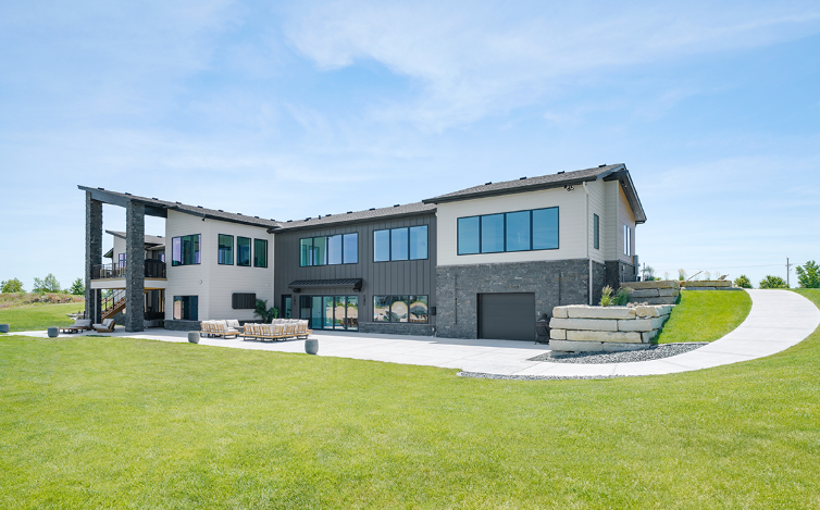 Modern two-story house with gray and tan siding, a long driveway, and a green lawn under a blue sky.