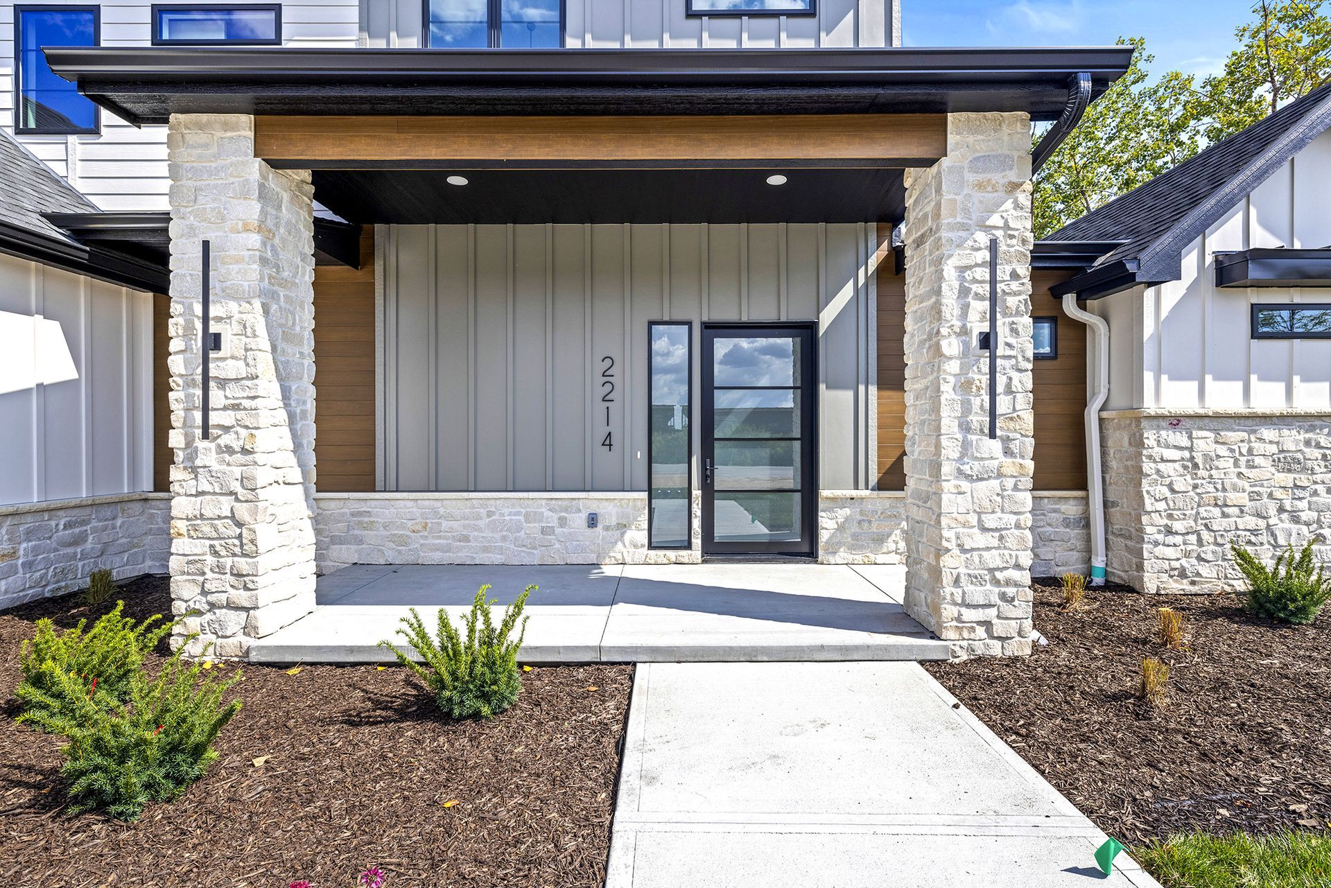 Modern front entrance with a glass door, light stone pillars, dark metal siding, and a concrete walkway in a yard.