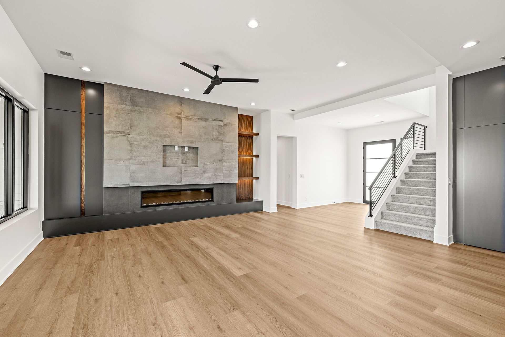 A modern living room with light wood floors, a large stone fireplace, and a staircase leading to a second floor.
