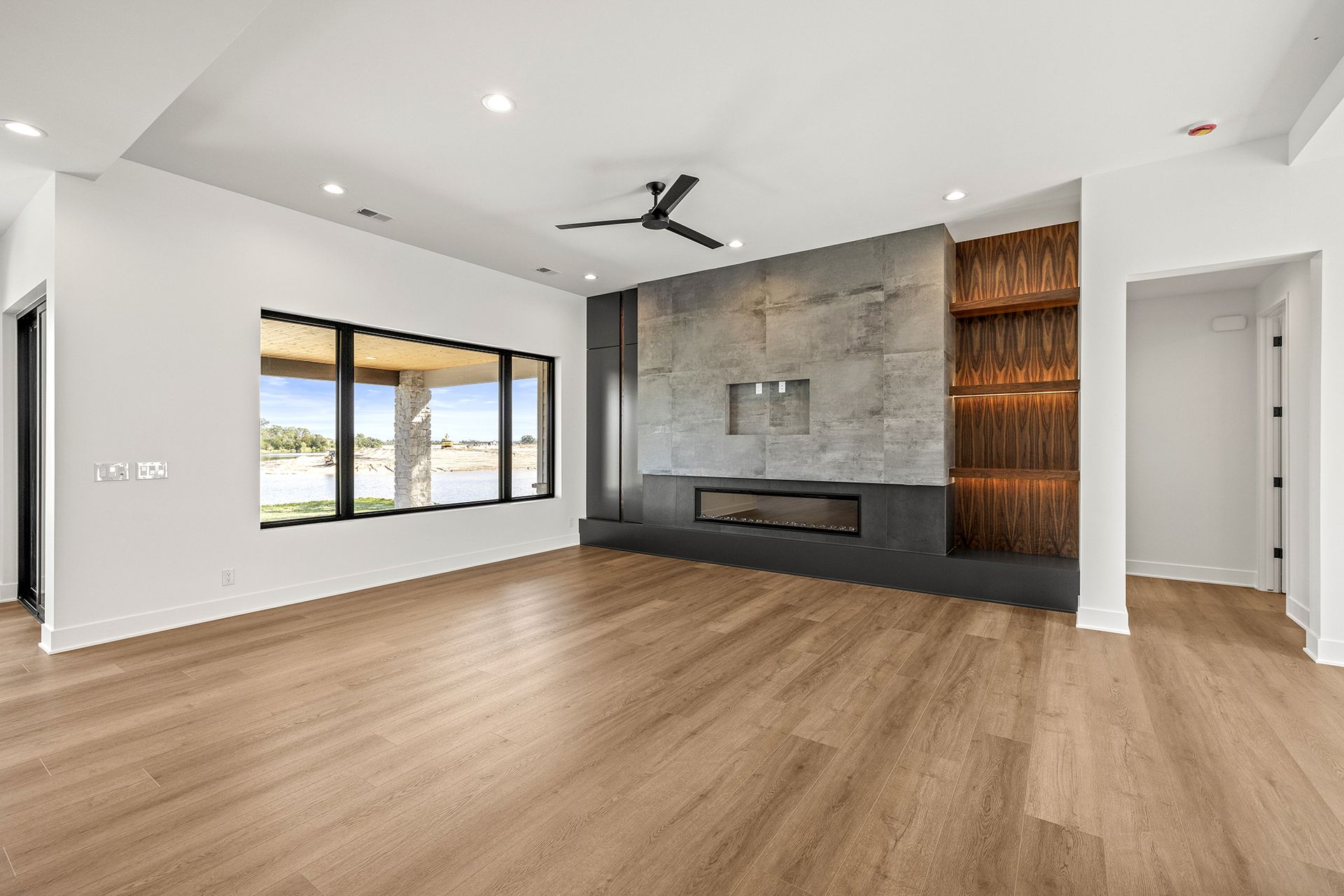 A bright, modern living room with light wood floors, a large picture window, and a gray stone fireplace with shelving.