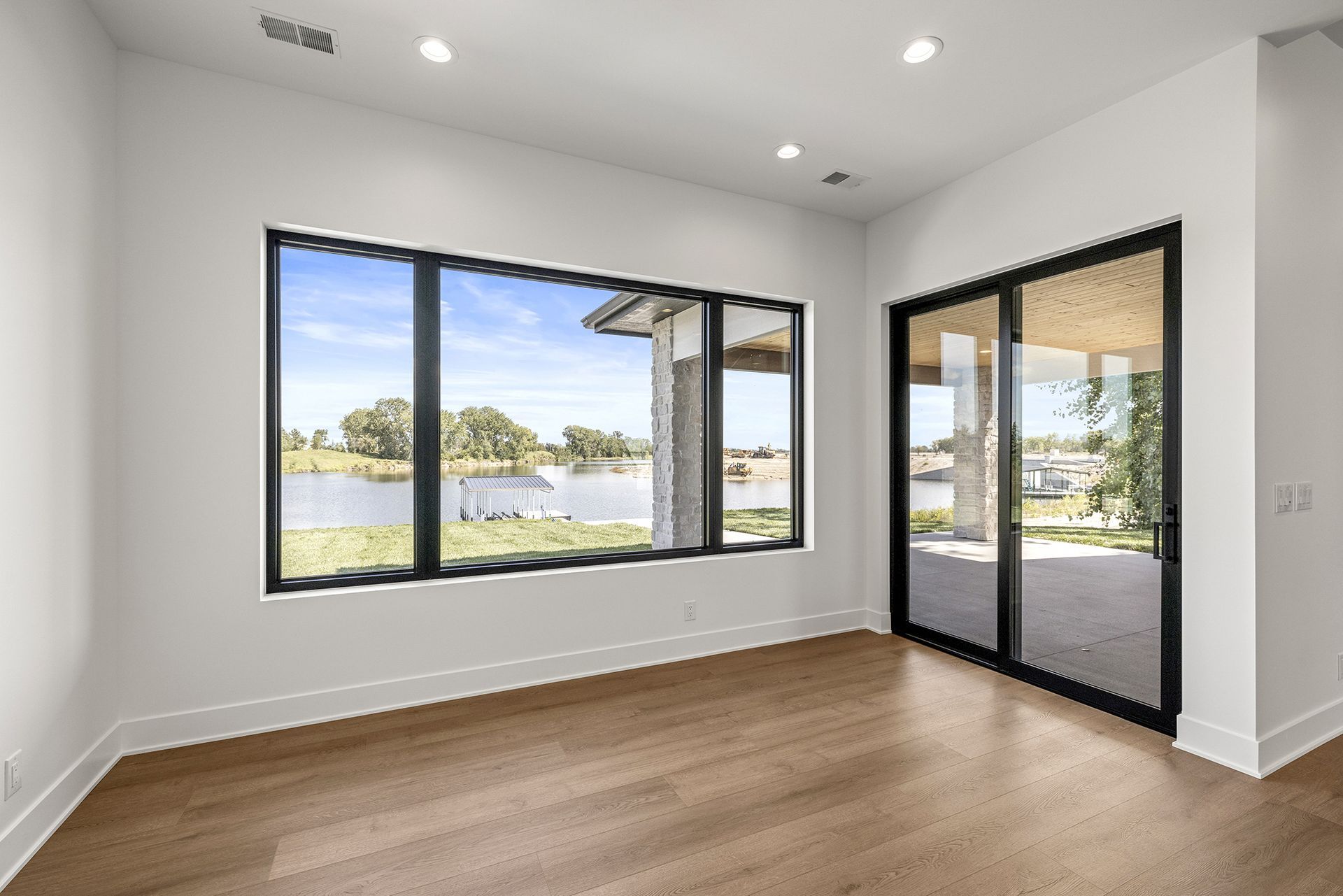 A bright, modern empty room with white walls, light wood floors, large black-framed windows, and a glass sliding door.