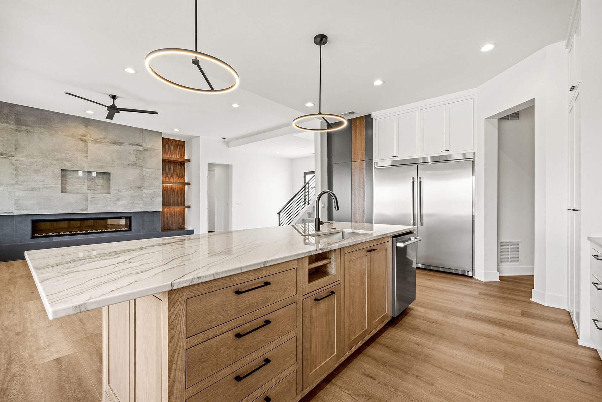 Modern kitchen with a light wood island, marble countertop, stainless steel refrigerator, and stone fireplace feature.