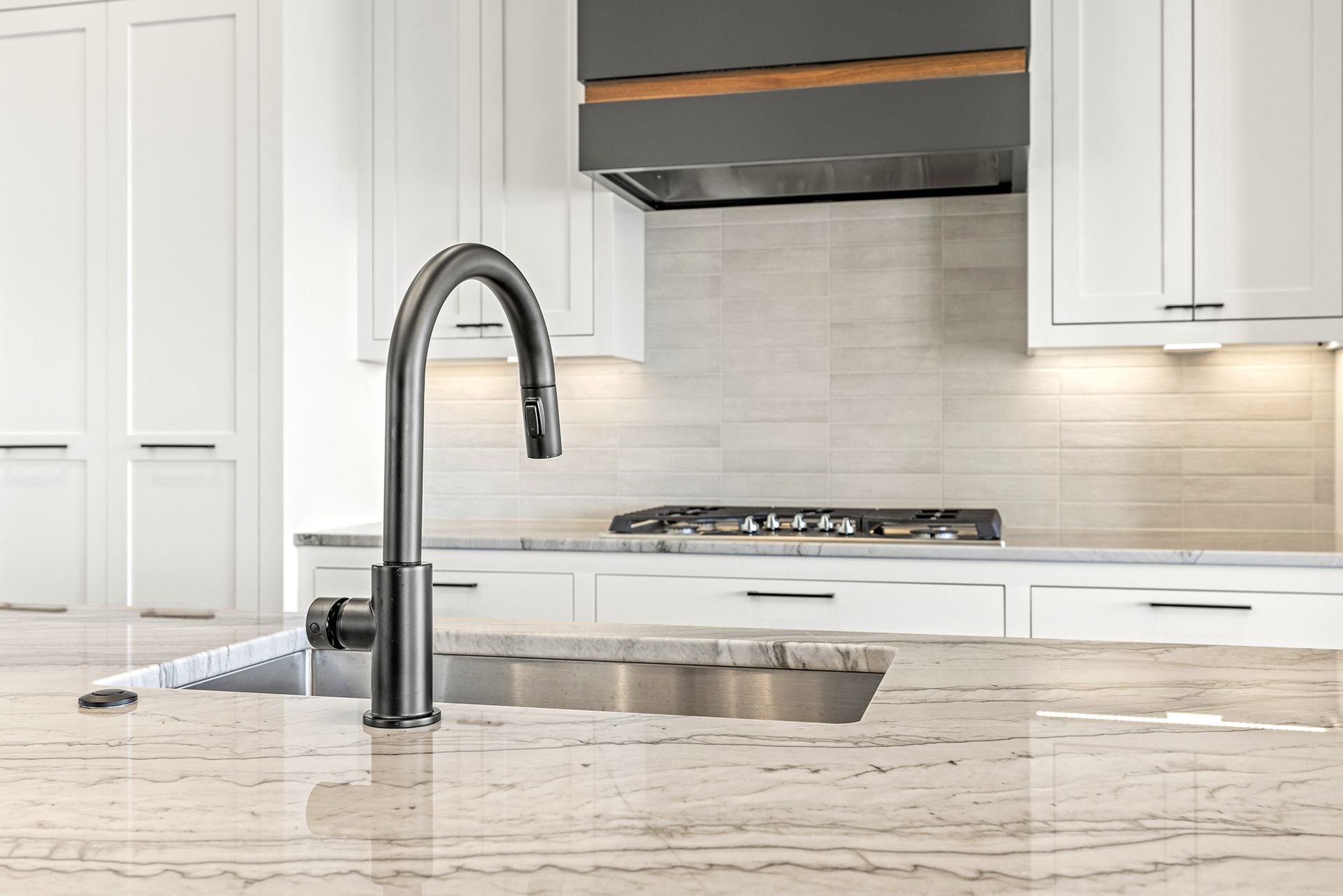 A dark metallic kitchen faucet stands on a marble countertop in front of white cabinets and a stovetop.