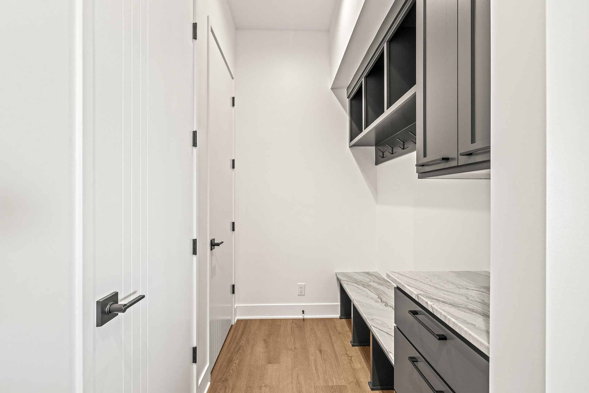 A modern mudroom with white walls, wood floors, dark grey cabinets, built-in bench seating, and a closed white door.