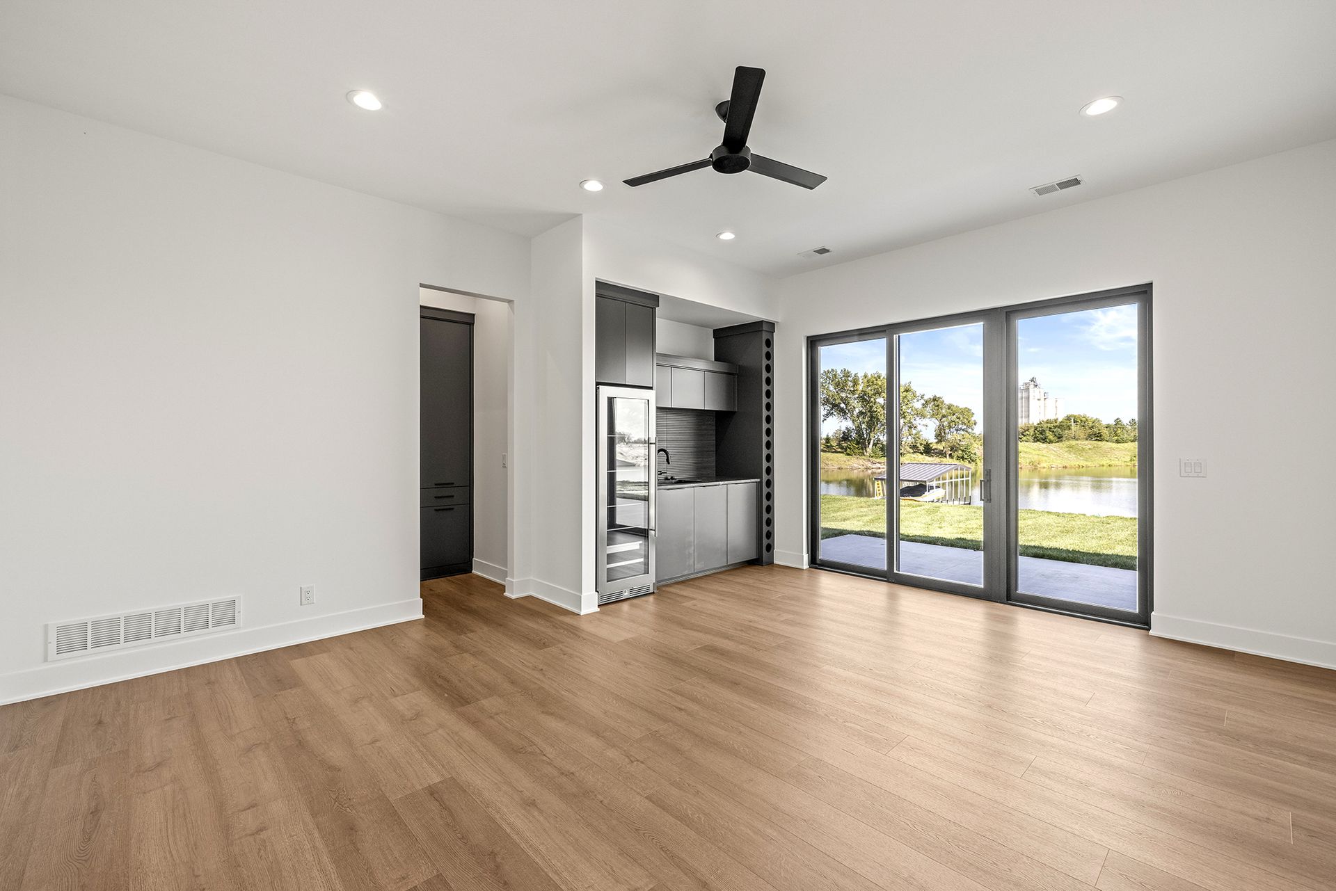 An empty modern room with light wood floors, a ceiling fan, a small built-in wet bar, and sliding glass doors to a pond.