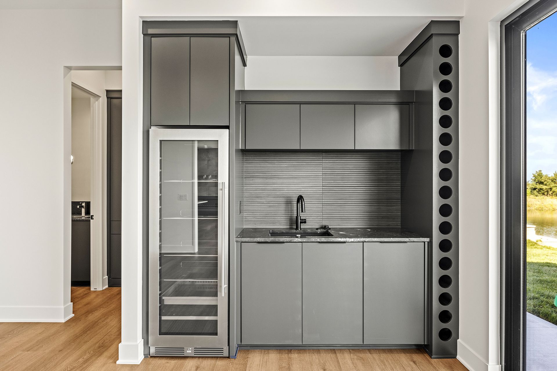 A modern home bar area with dark gray cabinets, a wine fridge, a sink, and a built-in vertical wine bottle rack.