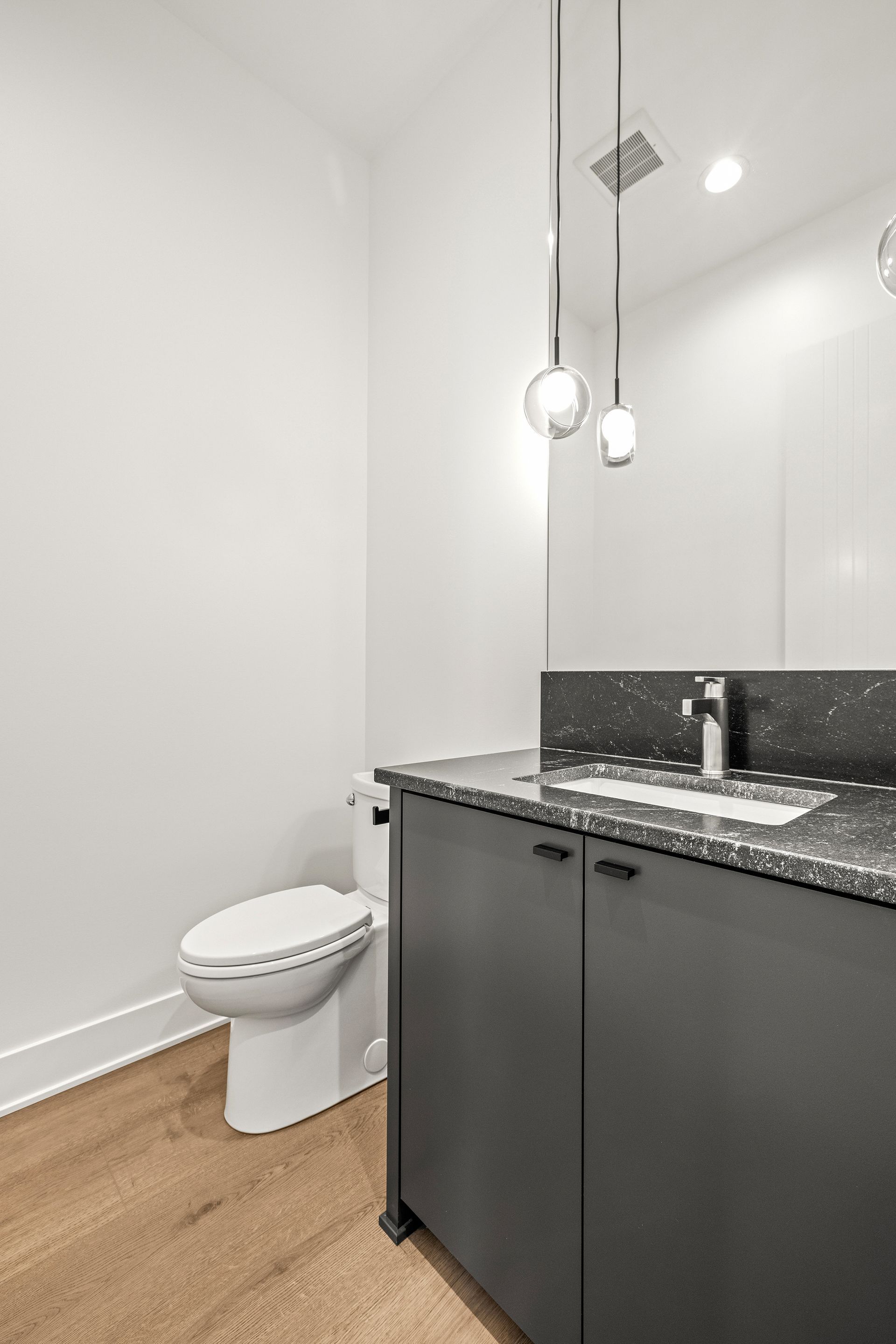 A modern bathroom with a charcoal vanity, speckled countertop, white toilet, light wood flooring, and hanging pendants.