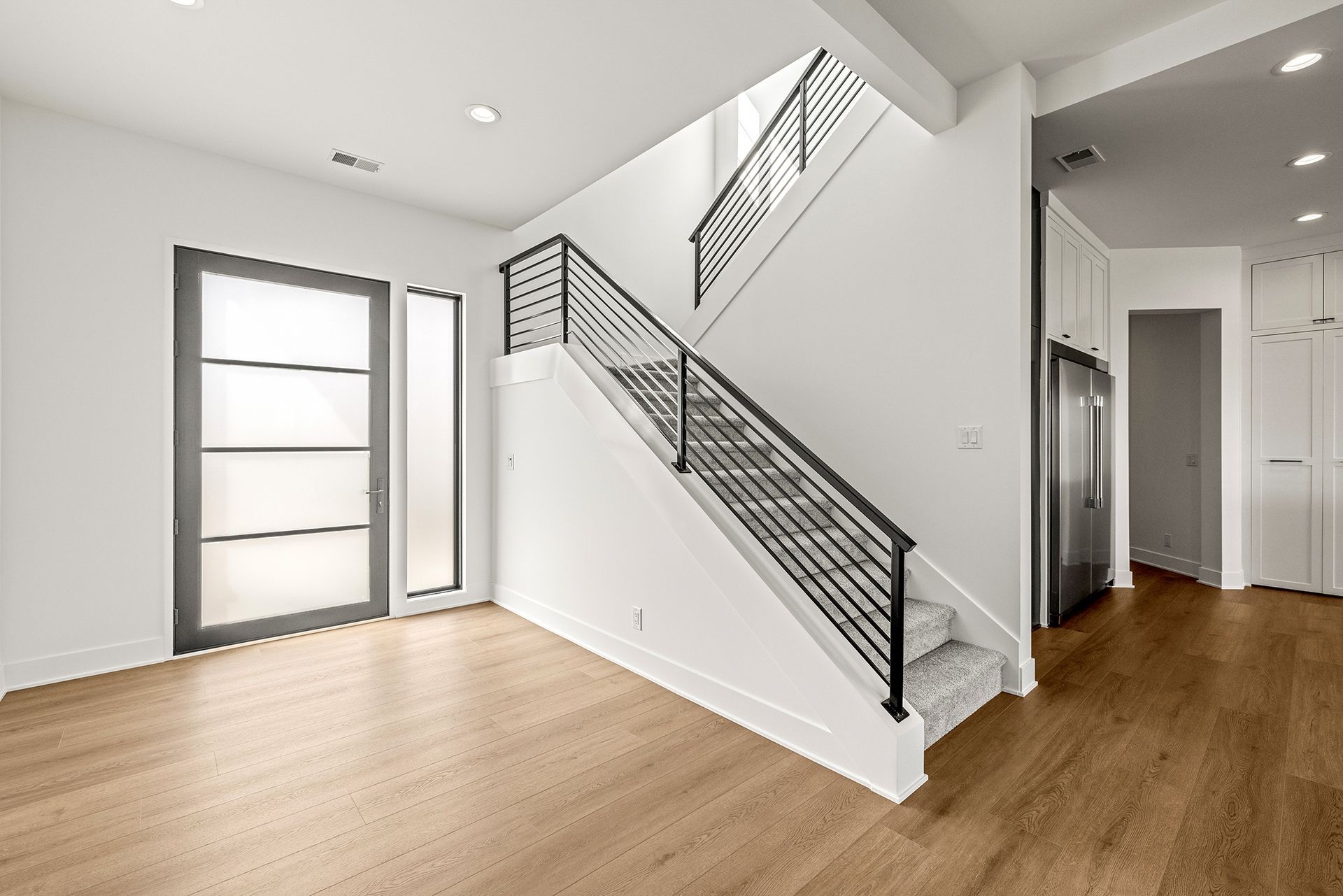 An interior view of a bright foyer with light wood floors, a modern glass front door, and a staircase with metal railings.