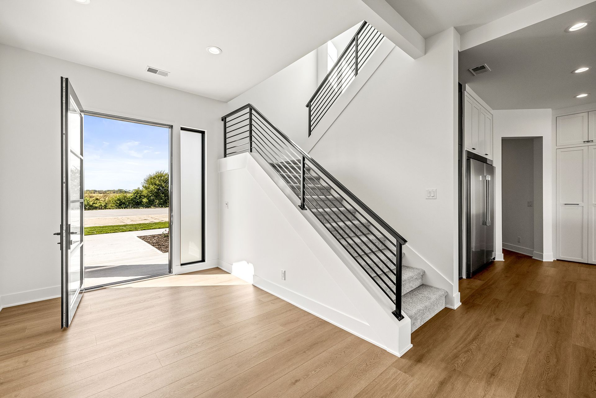A modern, white entryway featuring light wood floors, an open door, and a staircase with a black metal railing.