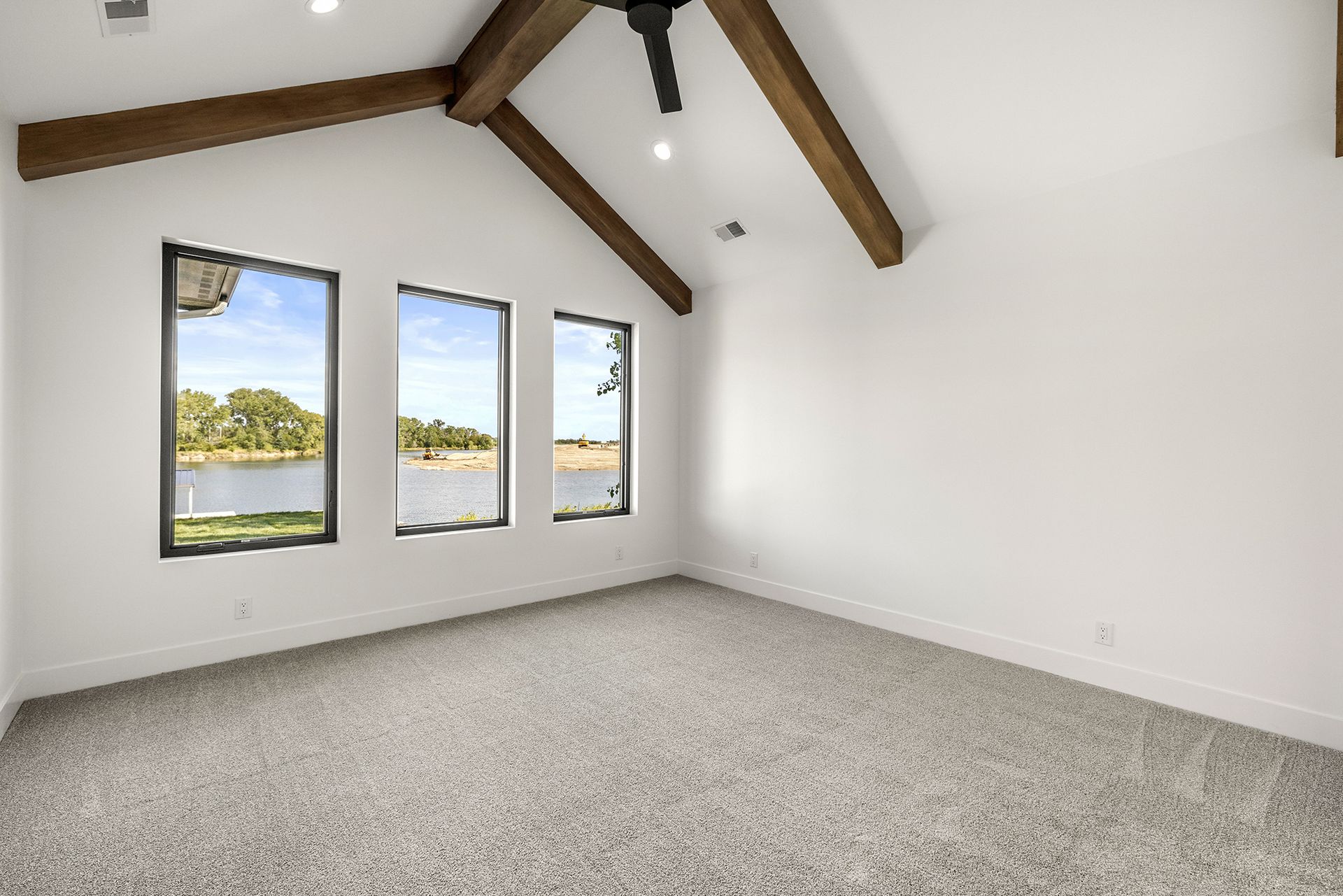 A modern, empty room featuring white walls, vaulted ceilings with wooden beams, gray carpet, and three windows facing water.