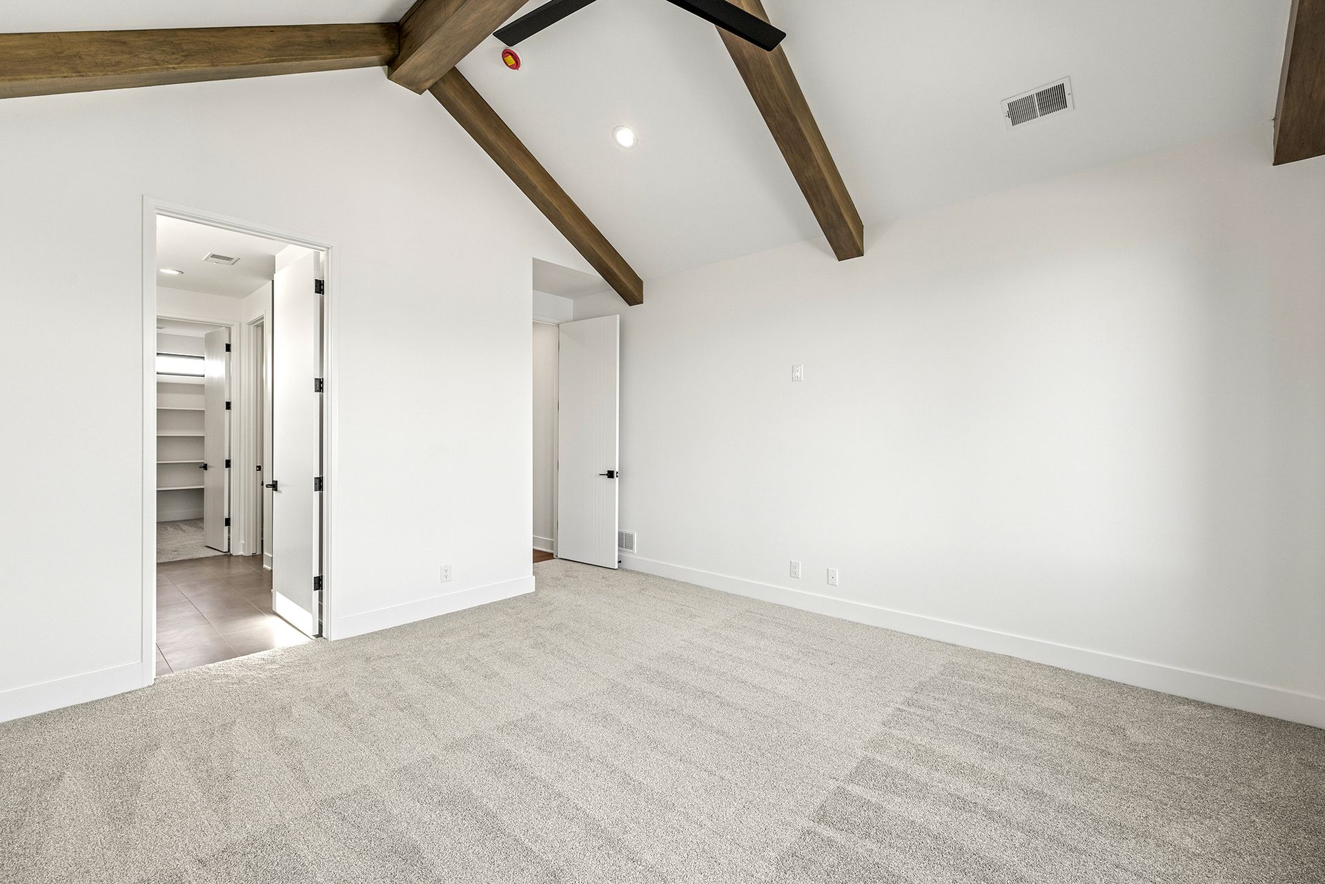 An empty bedroom with white walls, beige carpet, vaulted ceiling with wooden beams, and two open doorways.