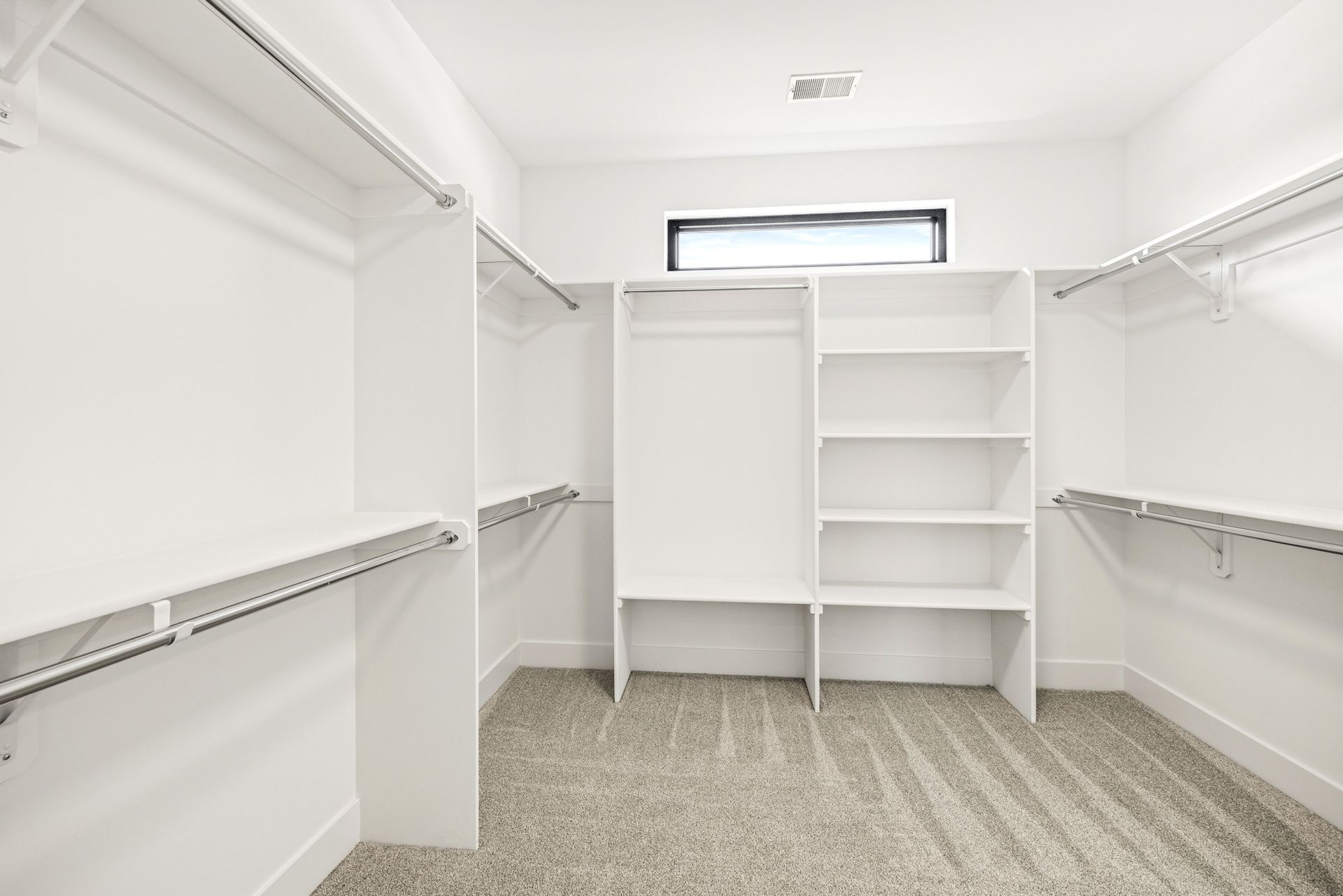 A walk-in closet with white shelving, hanging rods, and grey carpet under a horizontal window.