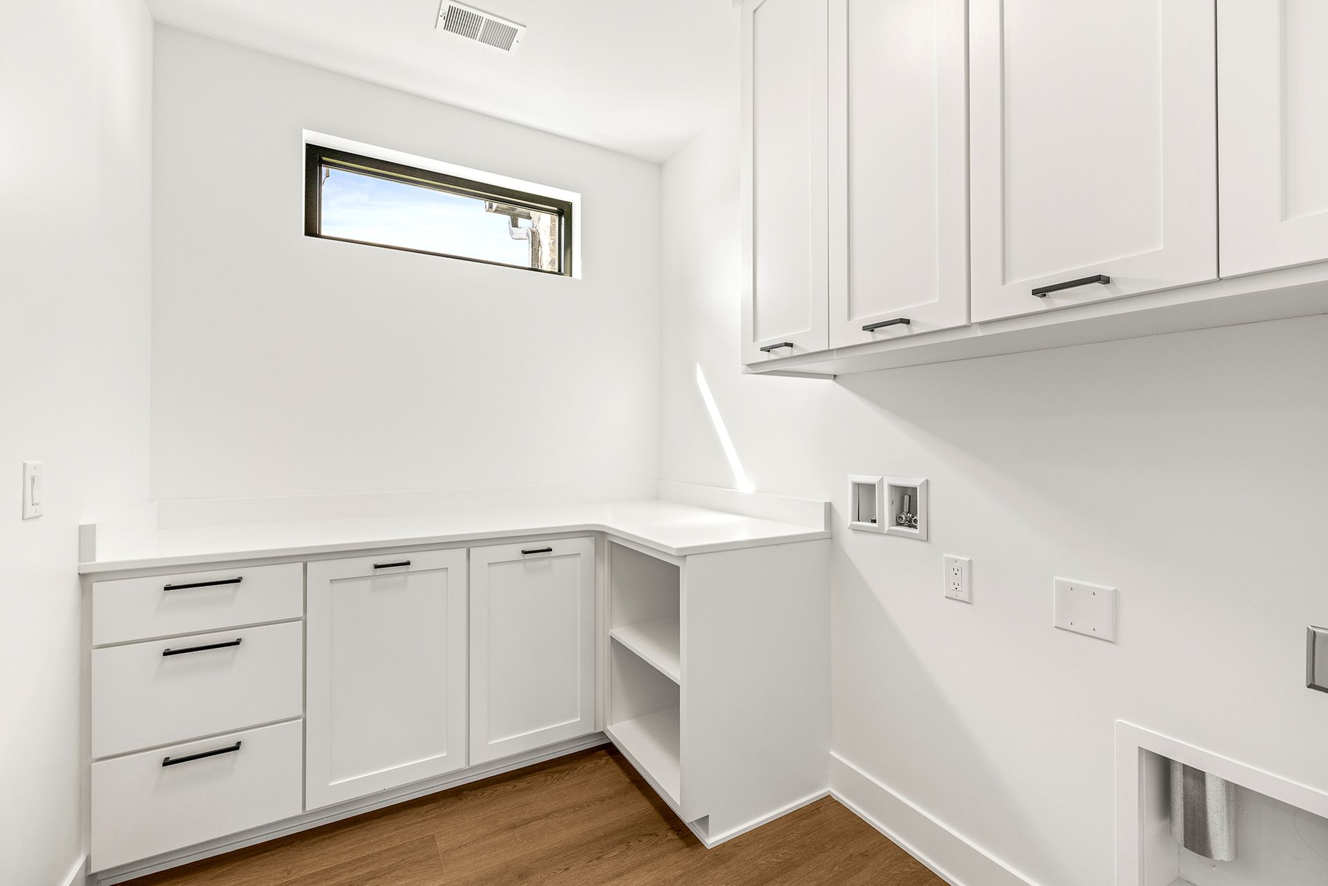 A modern laundry room with white cabinetry, a countertop, an open shelf, and a small window against a white wall.