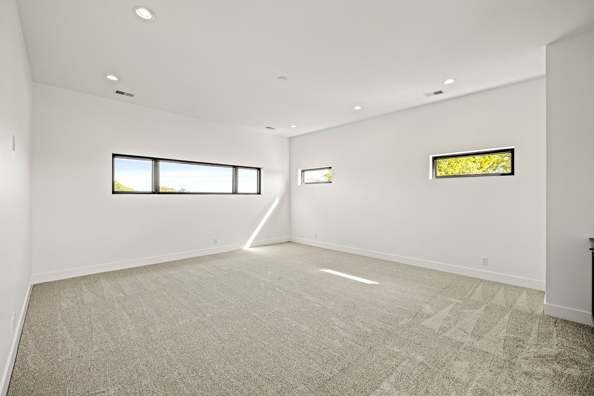 A bright, minimalist room with white walls, beige patterned carpet, recessed ceiling lights, and three horizontal windows.
