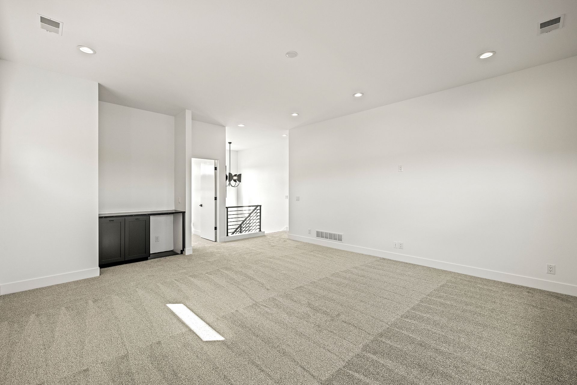 An empty room with white walls, beige carpet, recessed lighting, and a small built-in wet bar cabinet on the left.