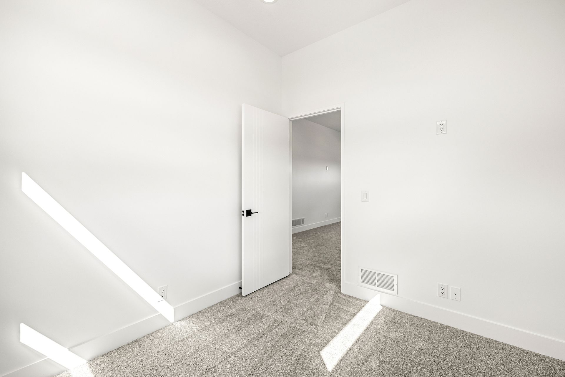 A bright, empty room with white walls, beige patterned carpeting, and an open white door leading to an adjacent space.