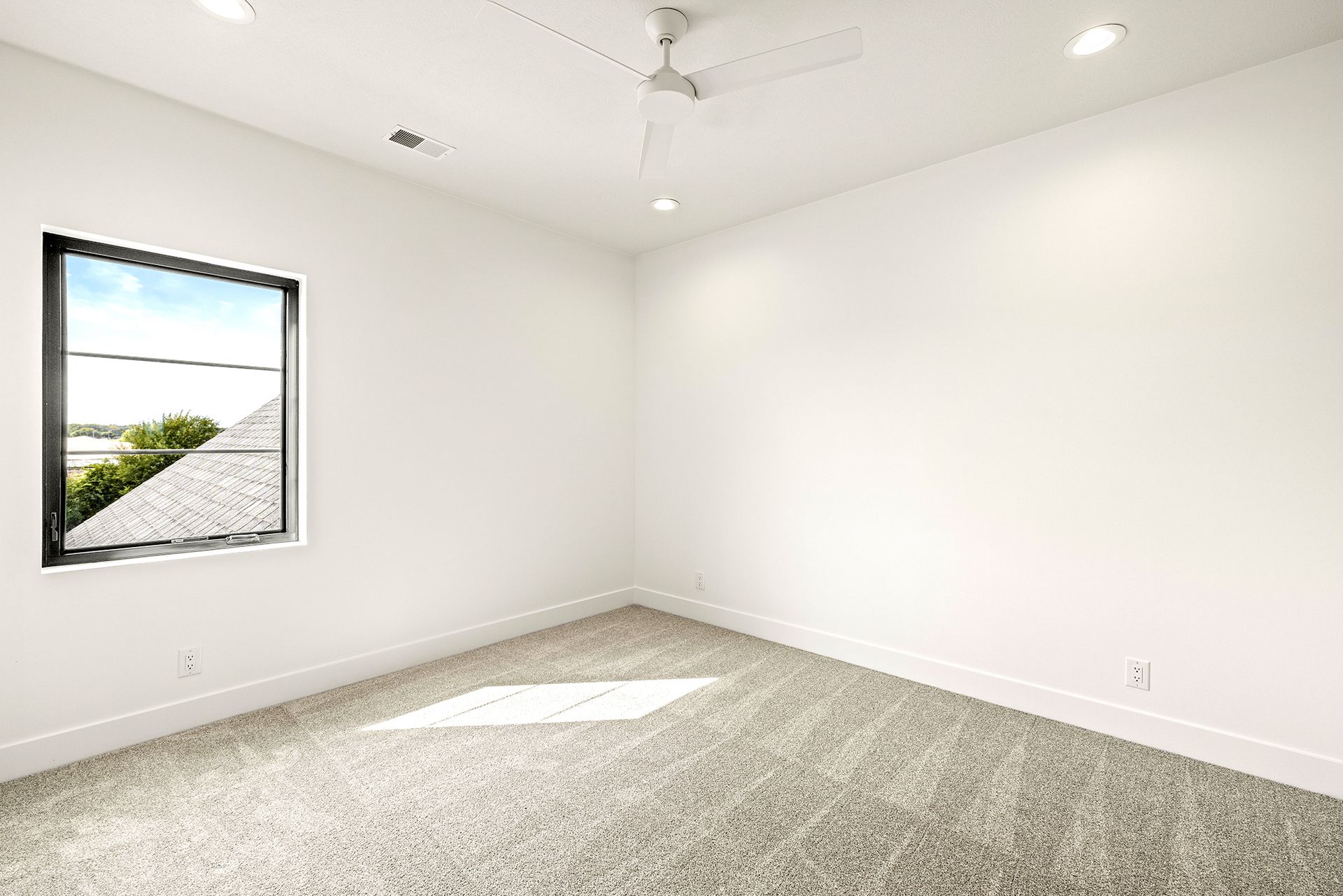 An empty room with white walls, gray patterned carpet, a ceiling fan, recessed lighting, and a square window.