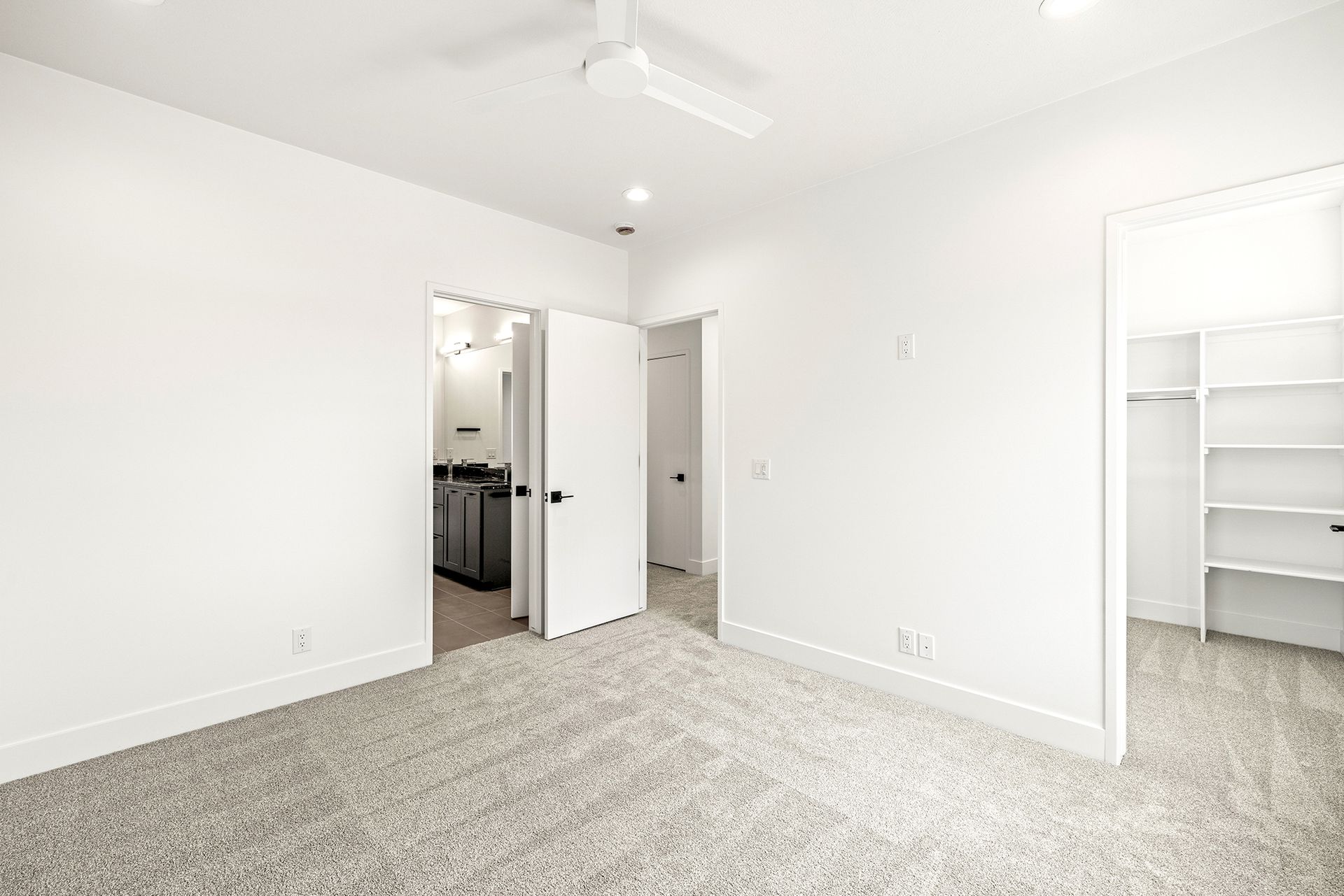 A minimalist, bright bedroom with white walls, gray patterned carpet, a ceiling fan, and open doorways to a closet and hall.